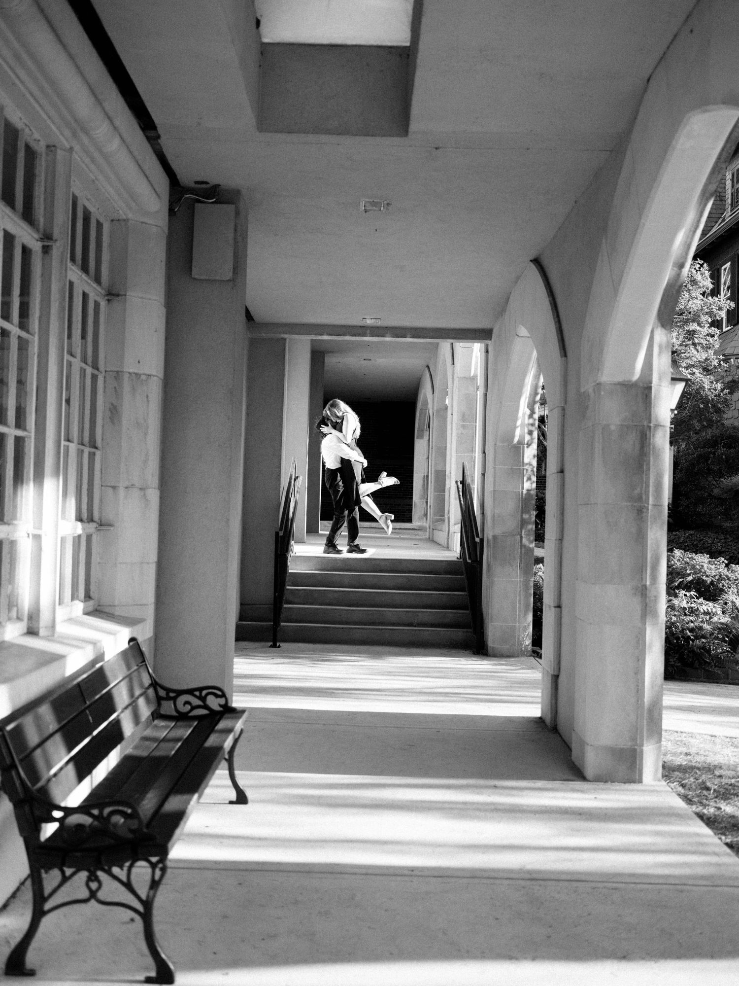Black and white photograph of an arched colonnade with sunlit walkway and empty bench at a historic building.