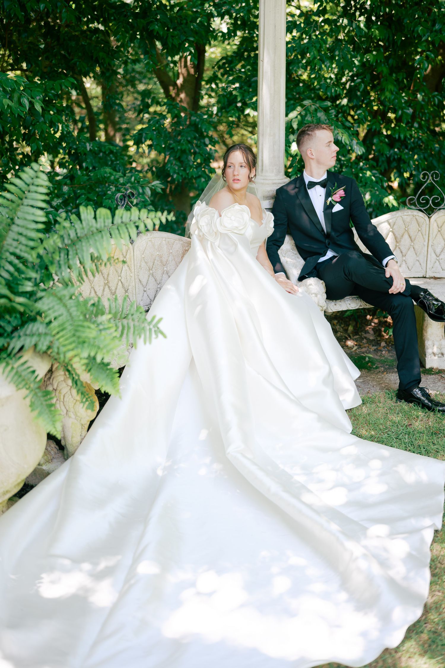 Bride in elegant white ballgown with long train sits beside groom in black tuxedo on stone bench in lush garden.