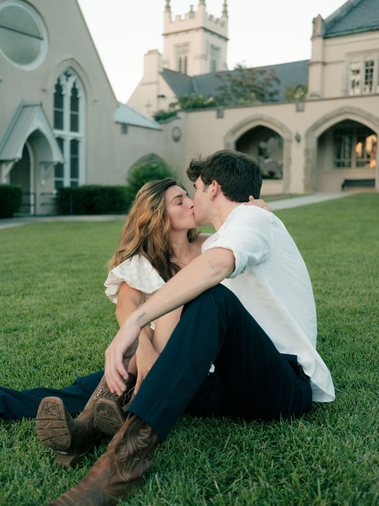 A romantic couple sitting on grass sharing a tender moment outside a beautiful gothic church with arched architecture.