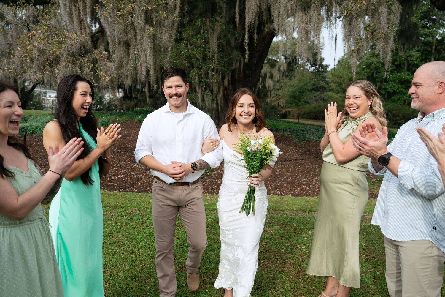 A joyful wedding party celebrates outdoors beneath Spanish moss-draped trees in a garden setting.