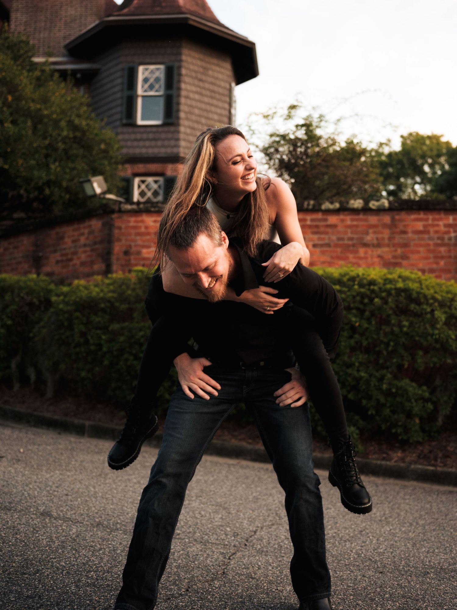 A playful couple share a piggyback ride moment outside a brick building with a turret at sunset.