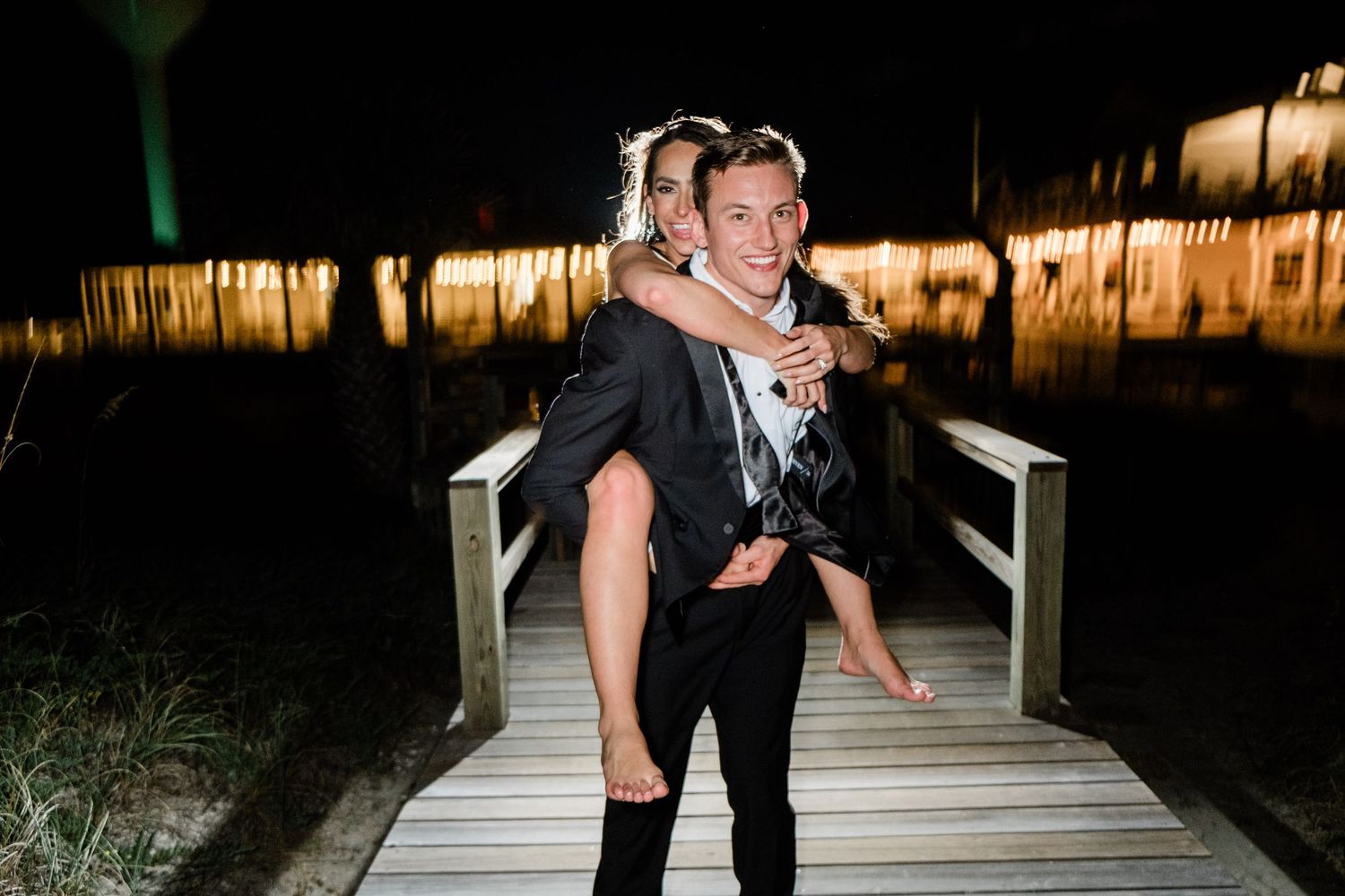 A couple in formal attire dances and poses together on a wooden deck at night with lights in the background.