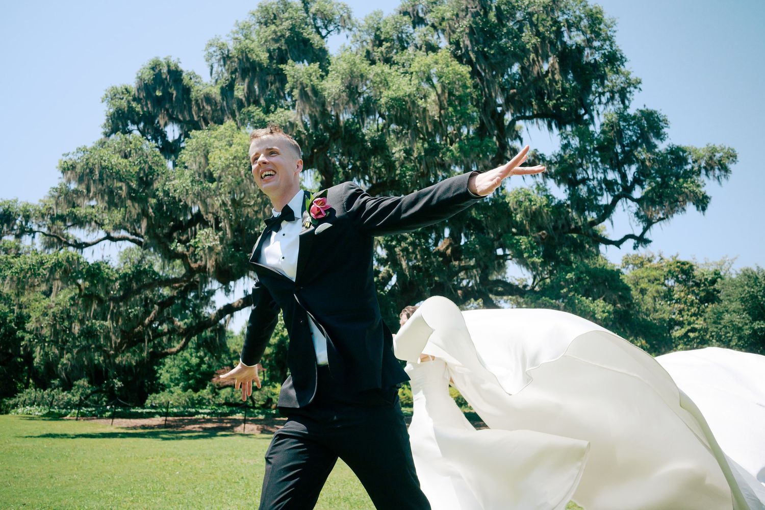 Groom in black tuxedo joyfully twirling bride in flowing white wedding dress outdoors near large oak tree.