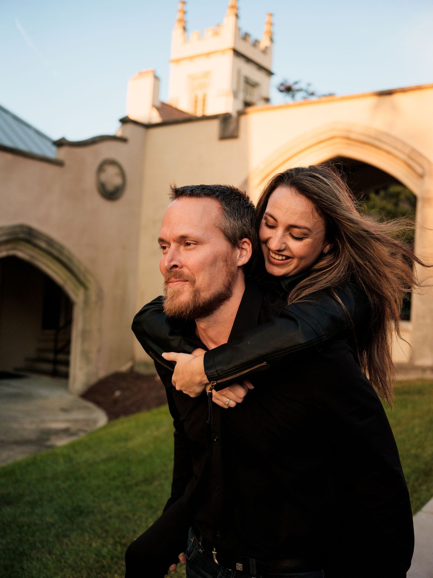 A couple shares a playful piggyback ride moment outside a historic church building at sunset.