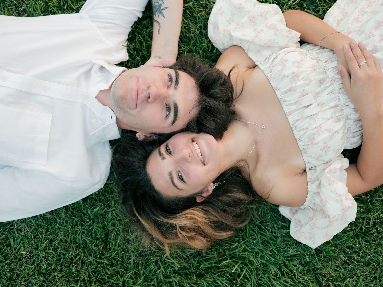A couple lies together on green grass wearing white clothing while relaxing outdoors.