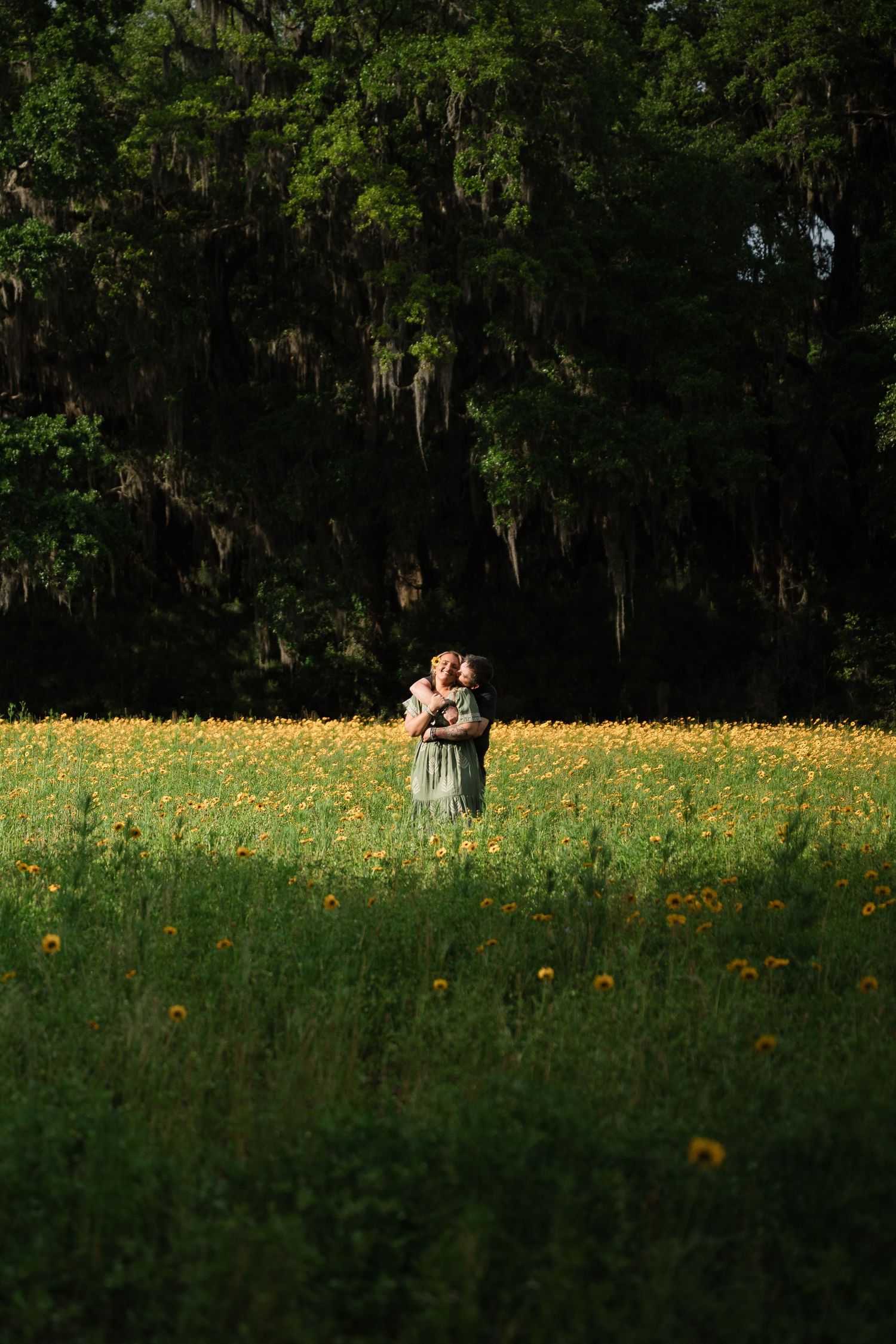 A couple sits together in a sunlit meadow filled with yellow wildflowers against a backdrop of dark forest trees.