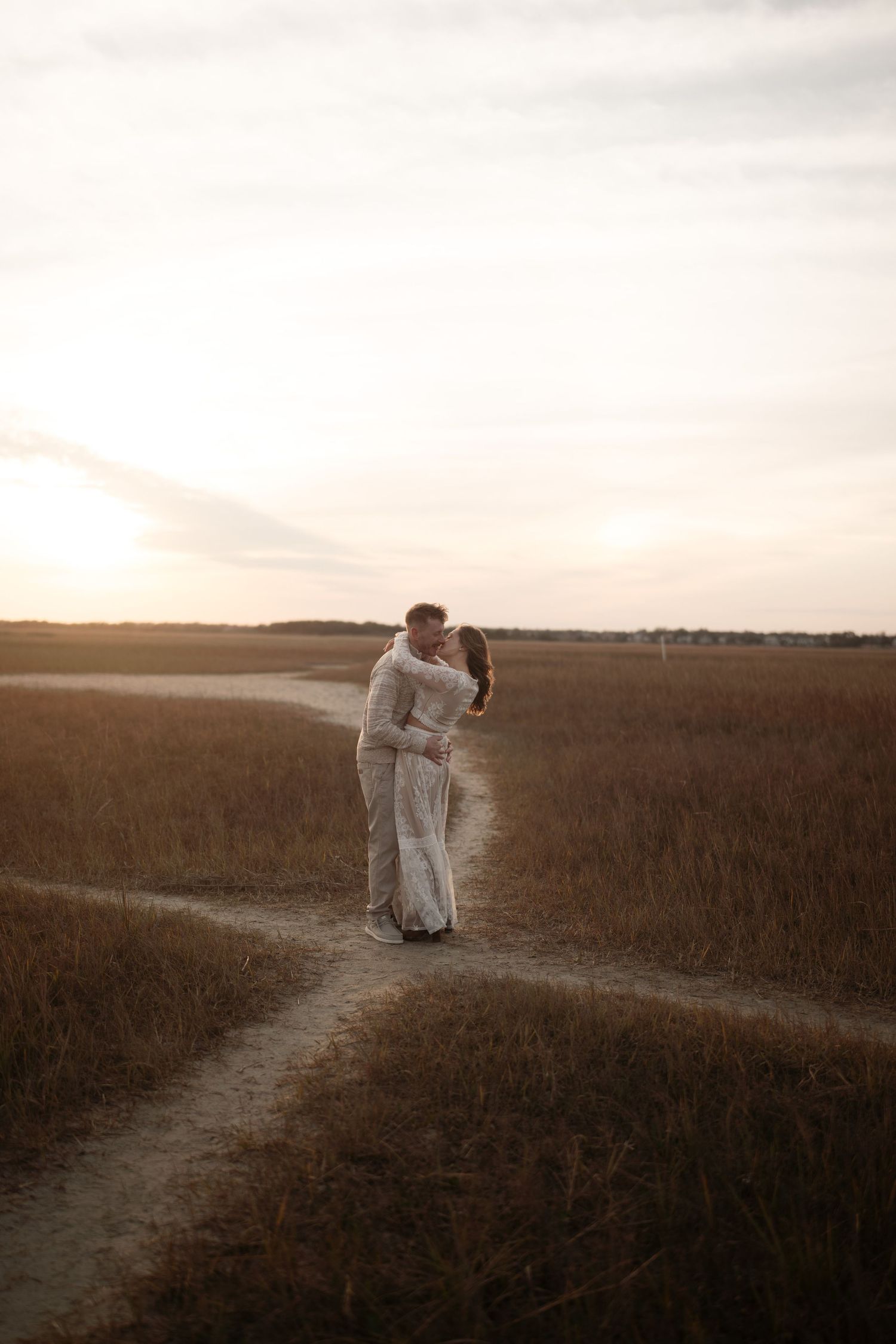 A couple embraces on a dirt path through tall grass during golden hour sunset in a serene rural landscape.