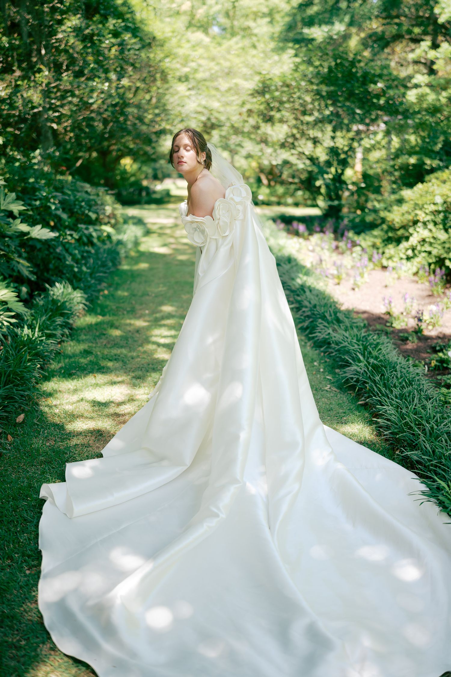 Bride in elegant white gown with long train stands in lush garden path, showcasing stunning backless wedding dress design.