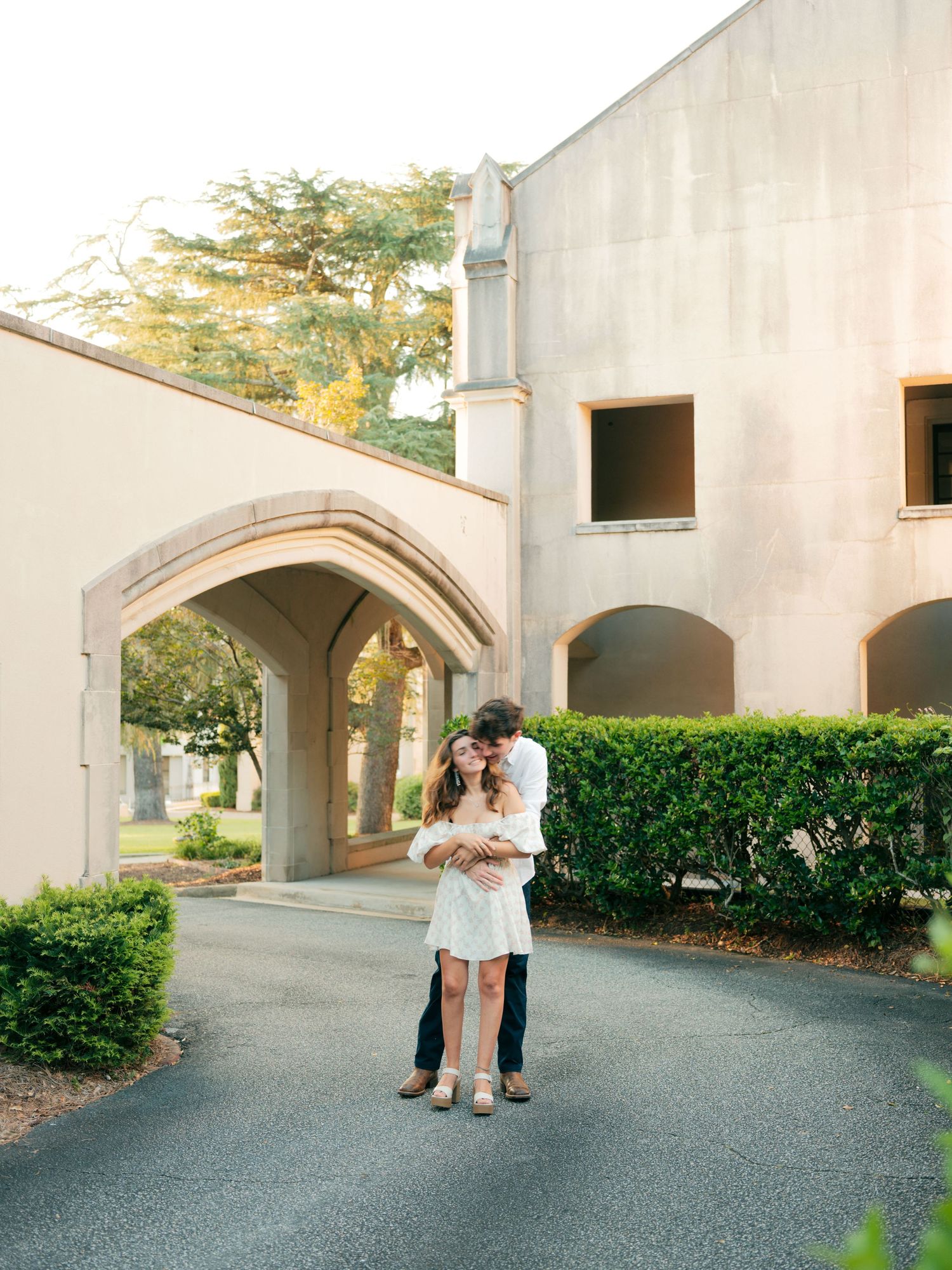 A romantic couple embraces on a paved path in front of an elegant arched building with manicured hedges.