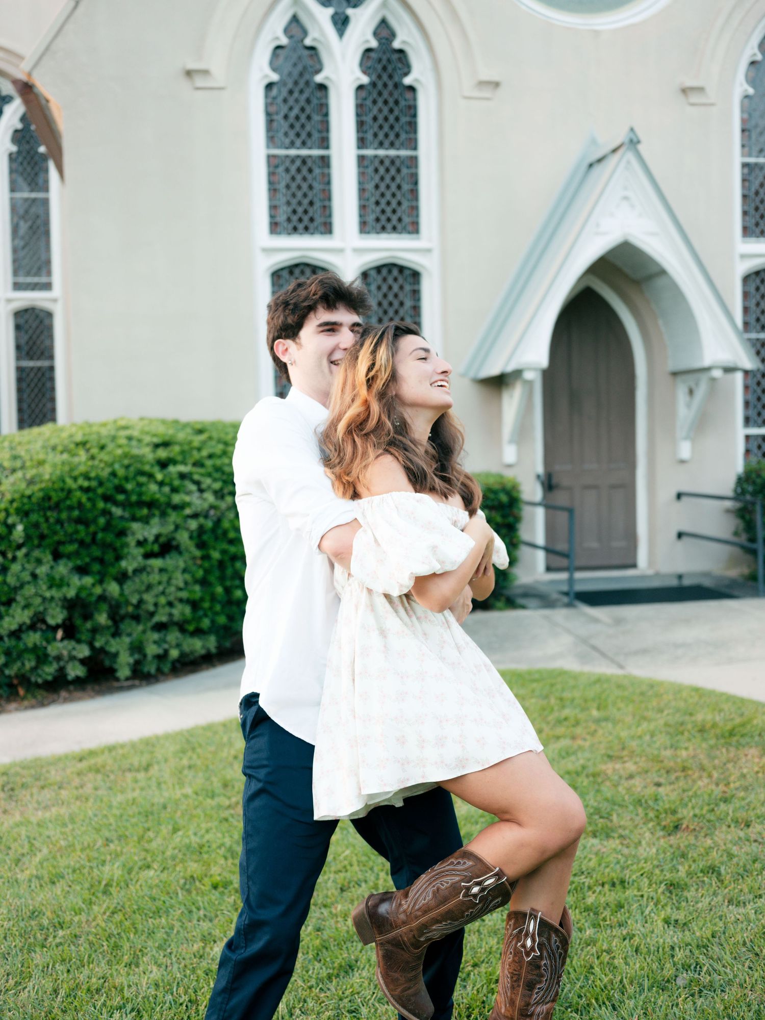A couple shares a joyful moment outside a gothic church with white walls and arched windows.