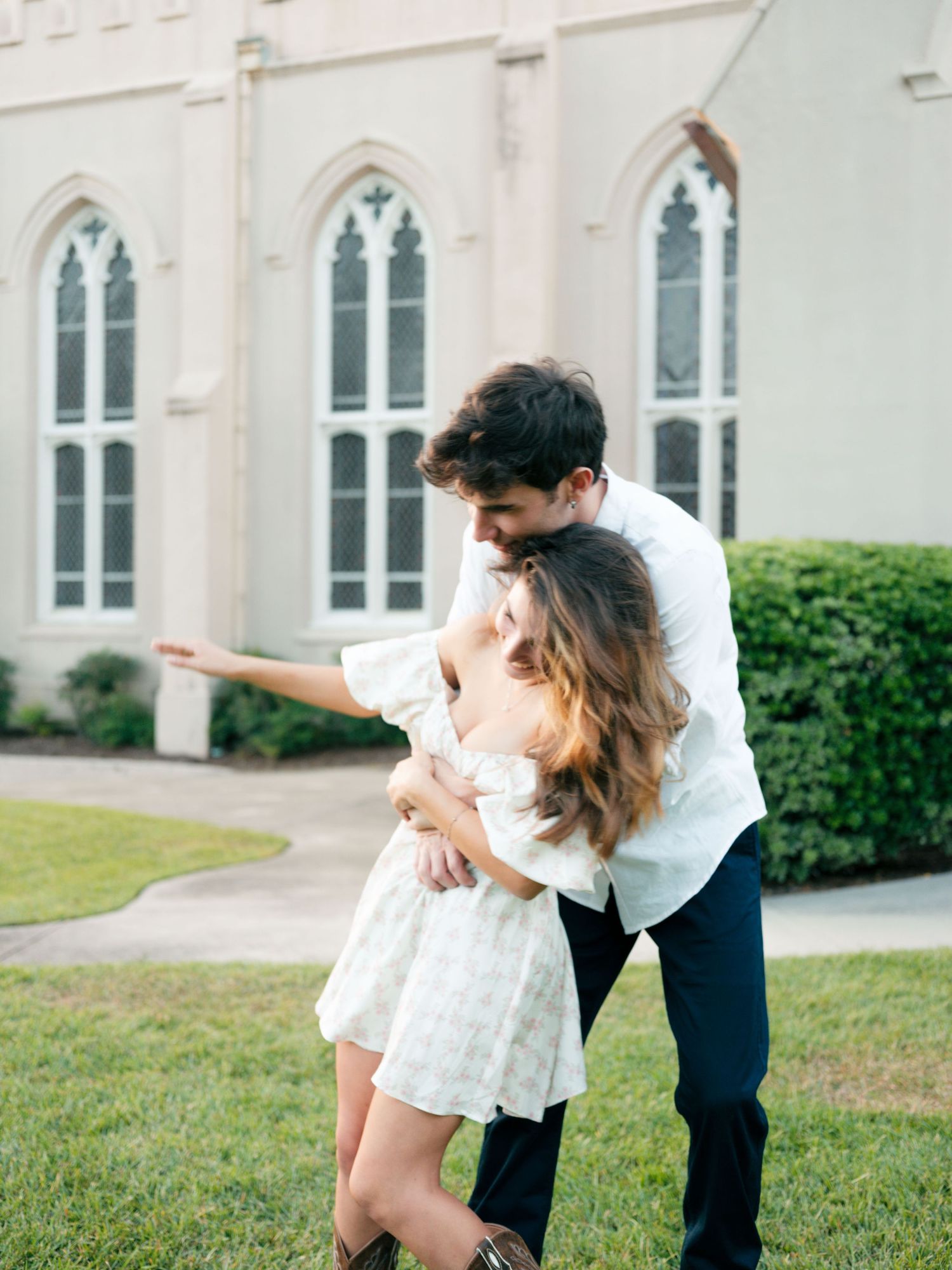 A playful couple dances together on the lawn in front of a historic church with Gothic architecture.