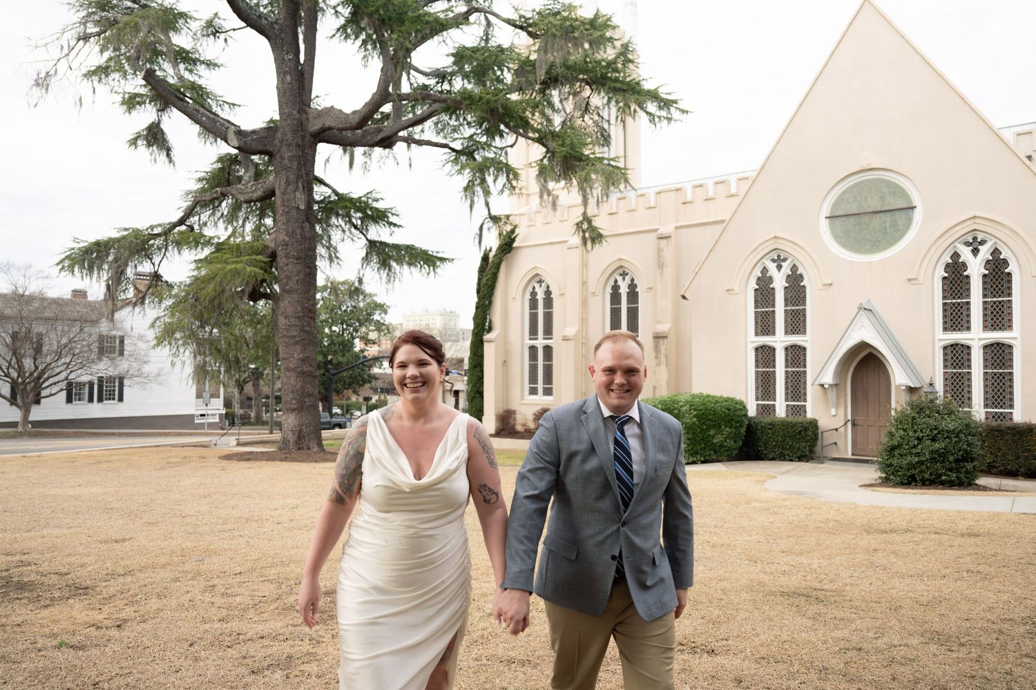 A couple holds hands while walking across the lawn in front of a white gothic-style church building.