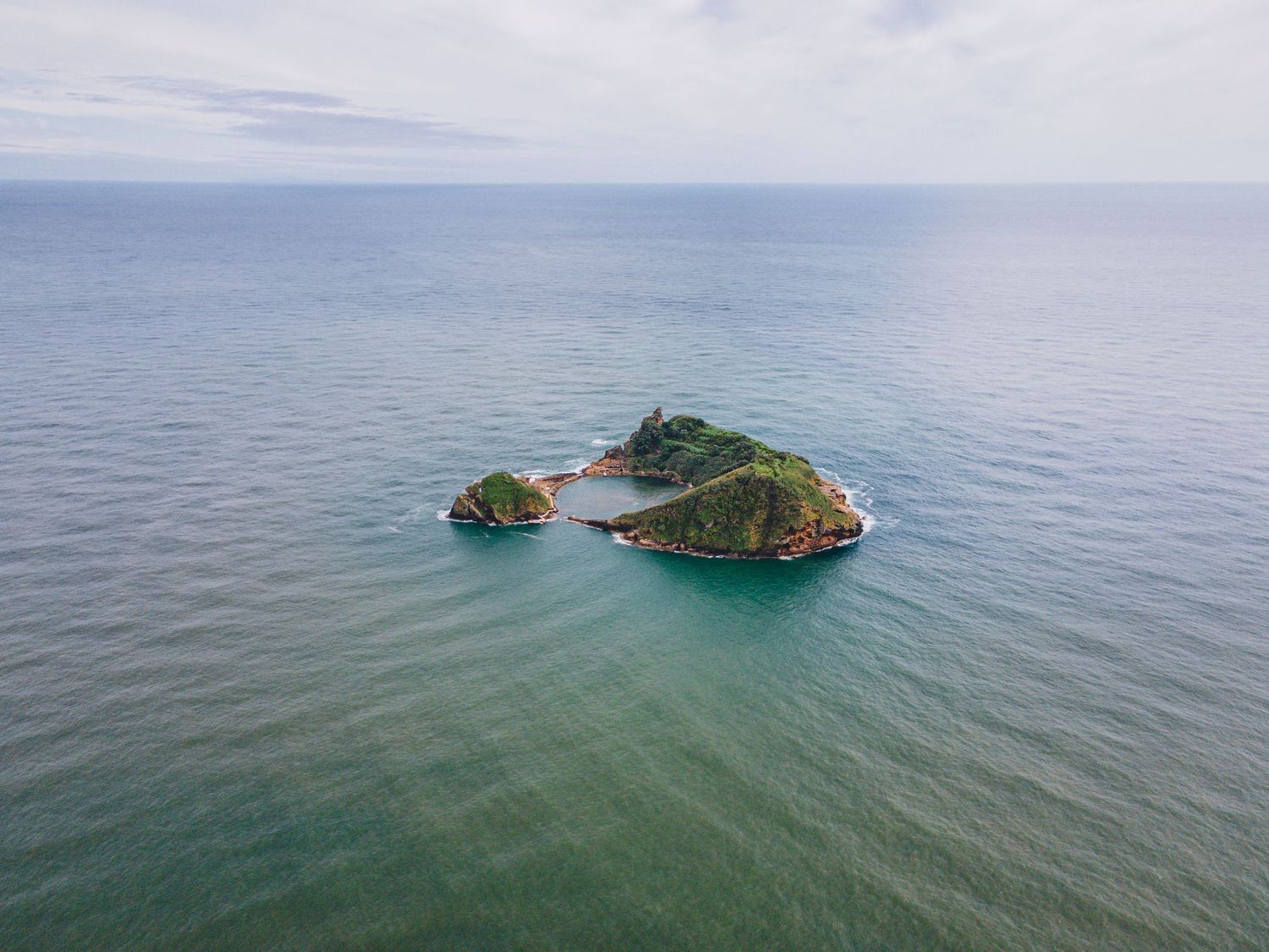 Aerial view of a small rocky island surrounded by turquoise waters in a calm ocean under cloudy skies.