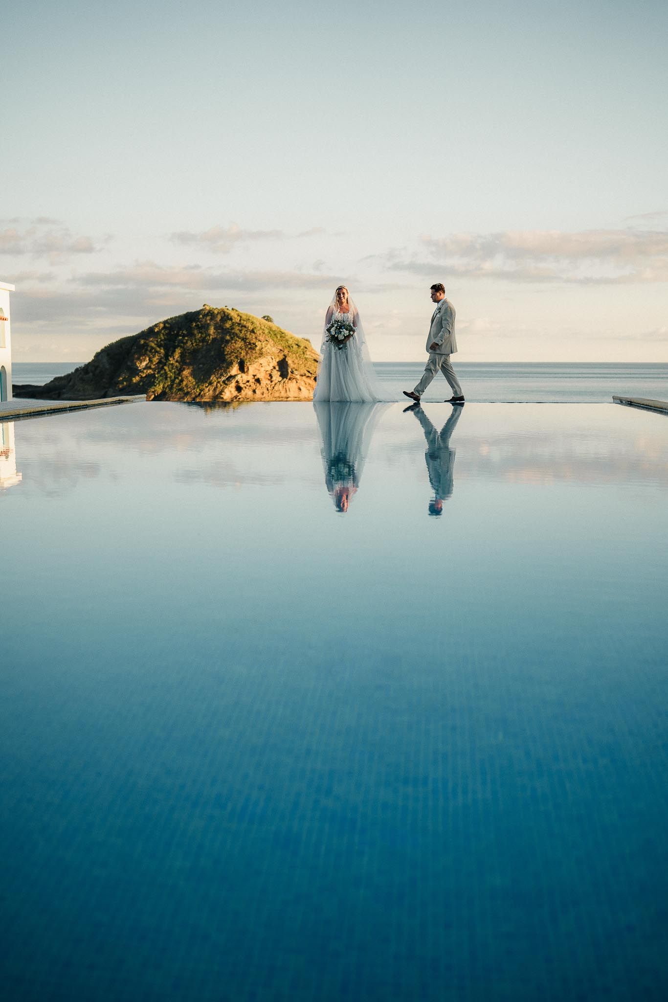 Two figures standing by an infinity pool at sunset with a rocky outcrop and ocean in the background.