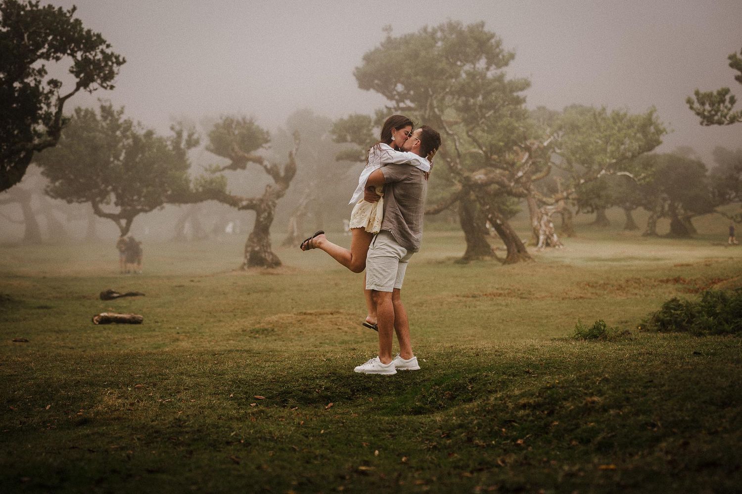 Two people embrace in a romantic moment on a misty hill surrounded by twisted trees on a foggy day at sunset.