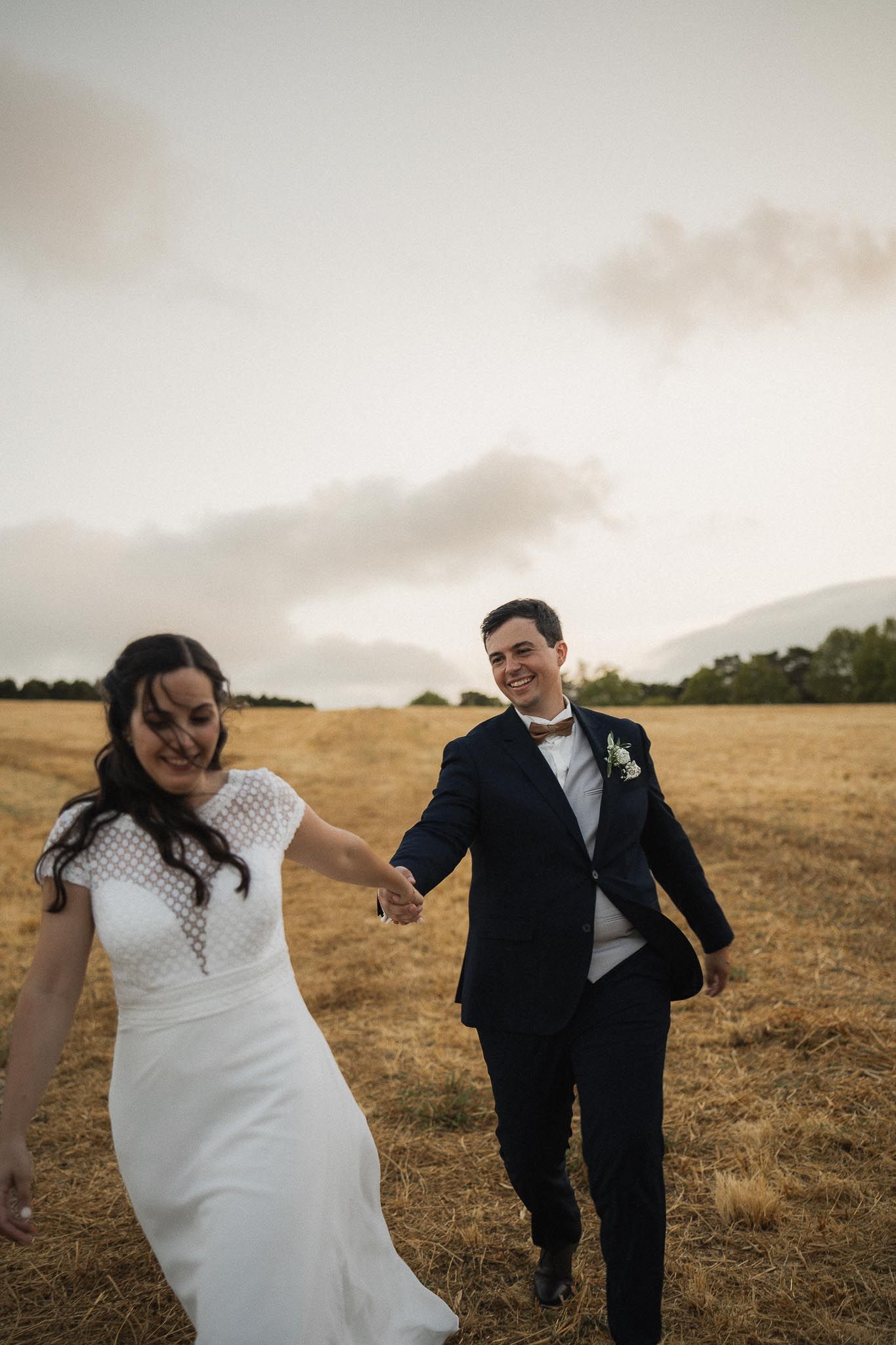 A couple in wedding attire dances playfully across a golden field at sunset.