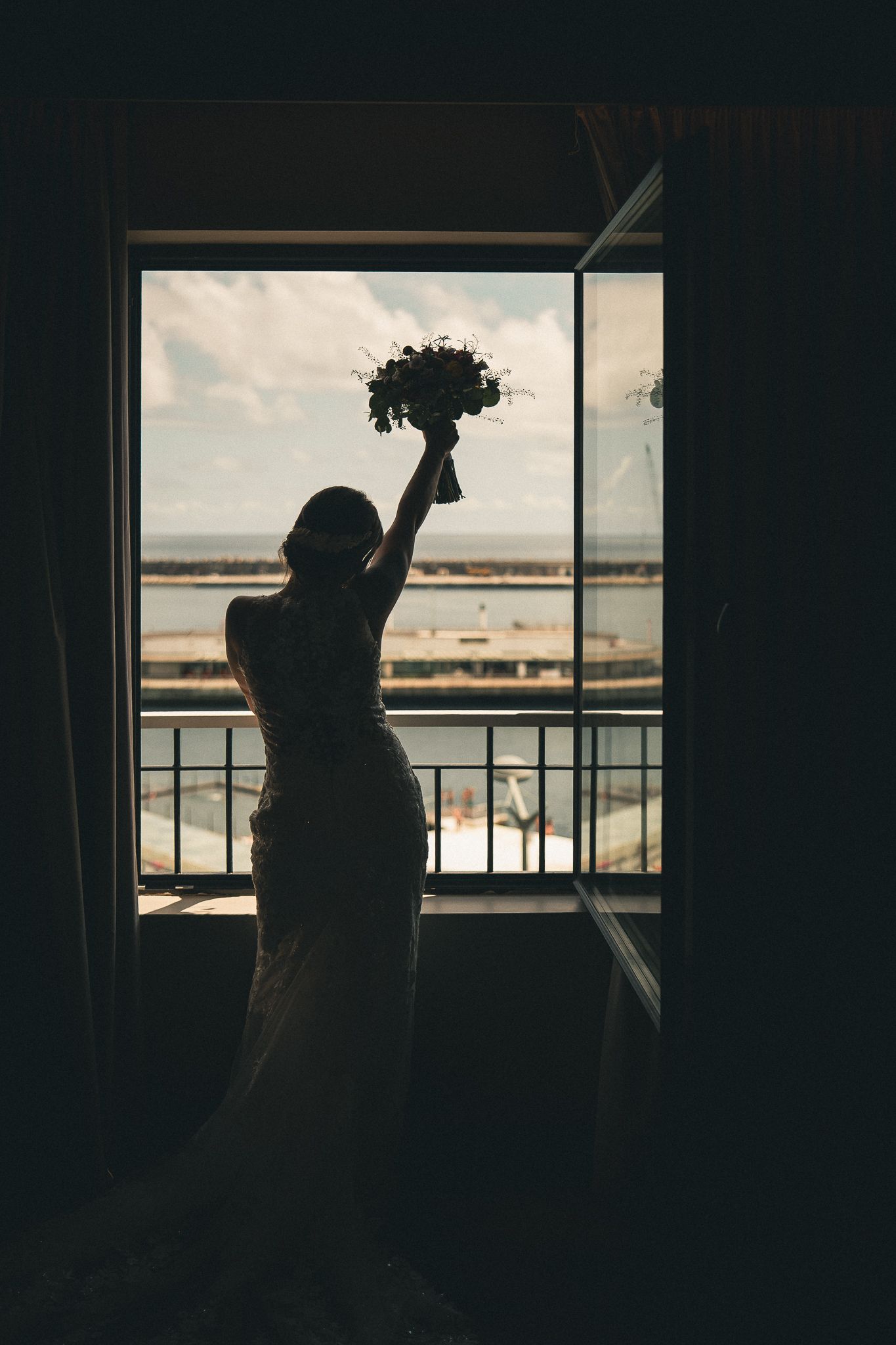 A silhouetted figure holds up a bouquet against a bright window overlooking a balcony at sunset.