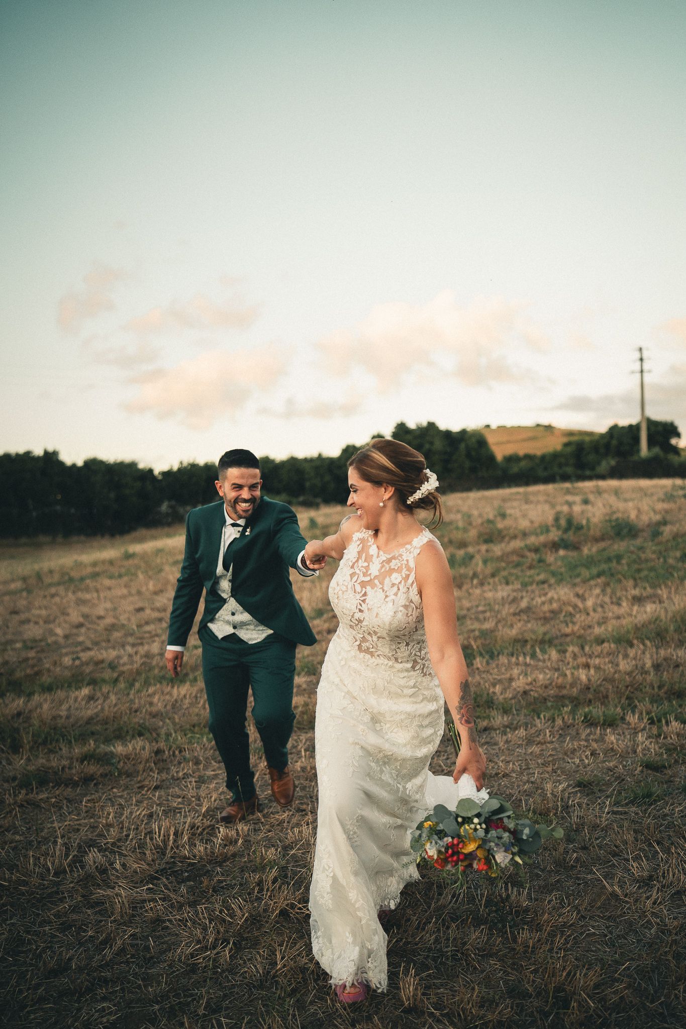 Couple in formal wedding attire walking together through a grassy hillside field at sunset.