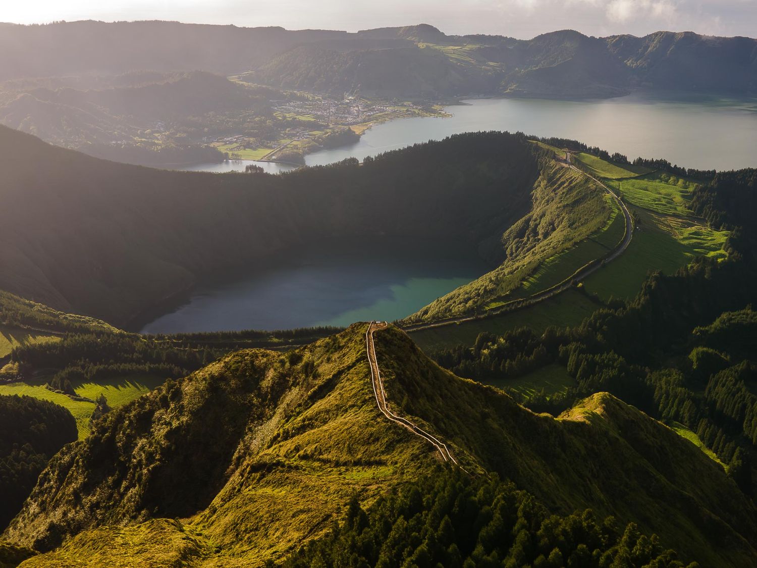 Golden sunlight illuminates rolling hills and volcanic lakes in a dramatic mountainous landscape.