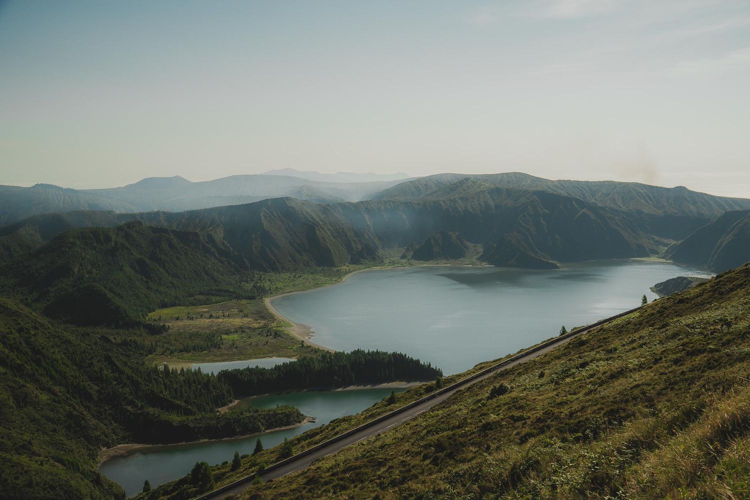 Aerial view of a stunning crater lake surrounded by green mountains and winding roads in a volcanic landscape.