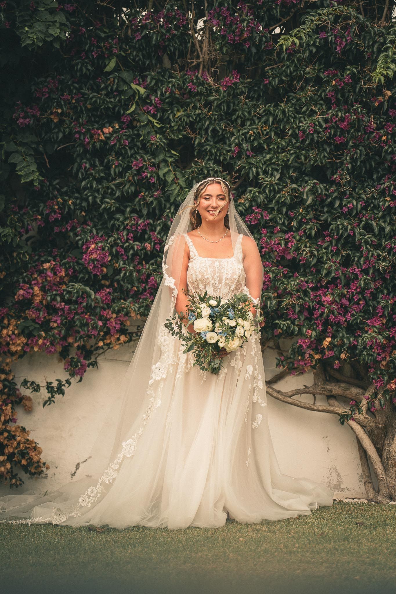A bride in a flowing white gown and veil stands before a wall of vibrant pink bougainvillea flowers holding a white bouquet.
