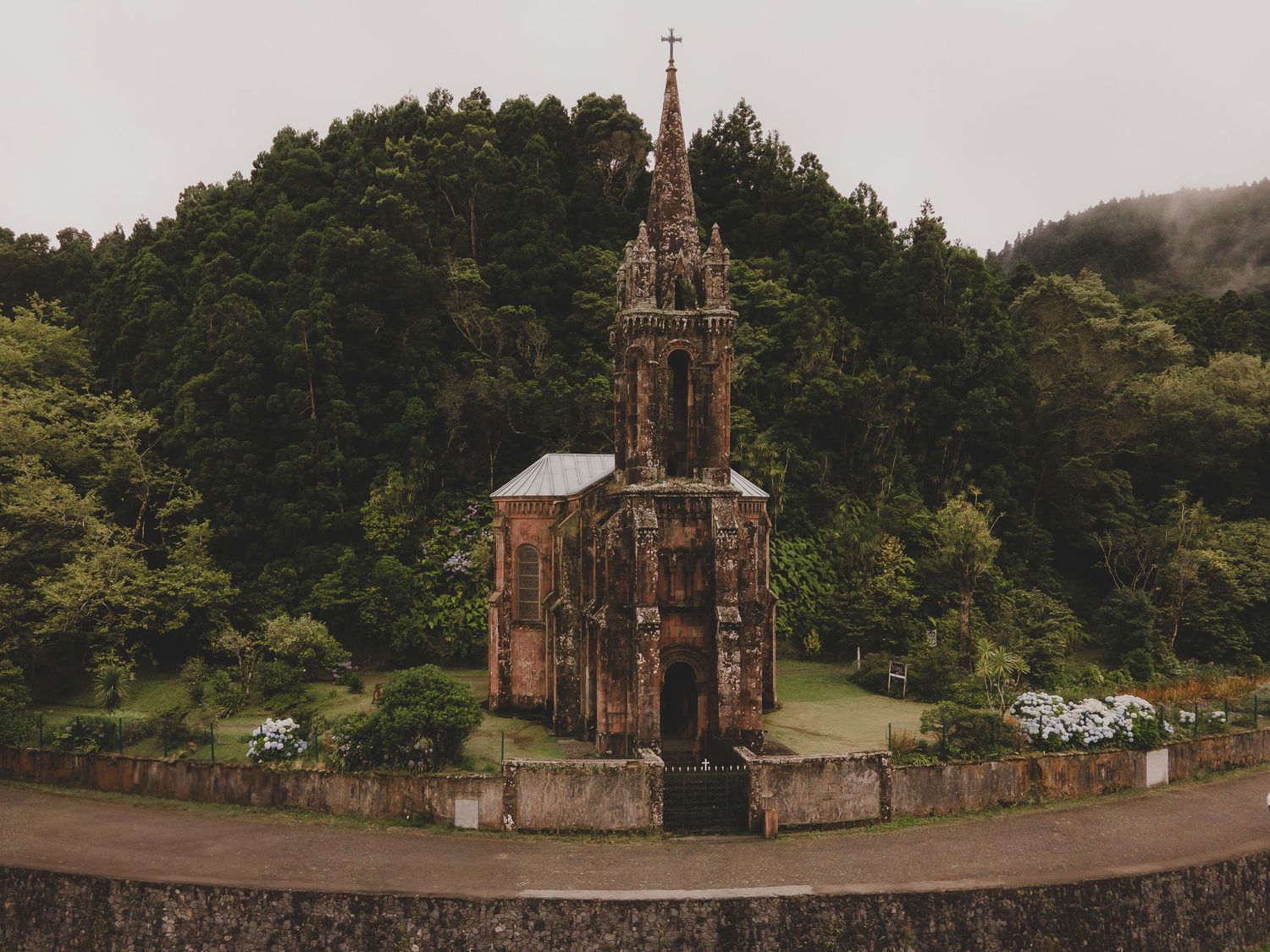 A Gothic revival church spire stands isolated among green forest hills in a tranquil rural setting.