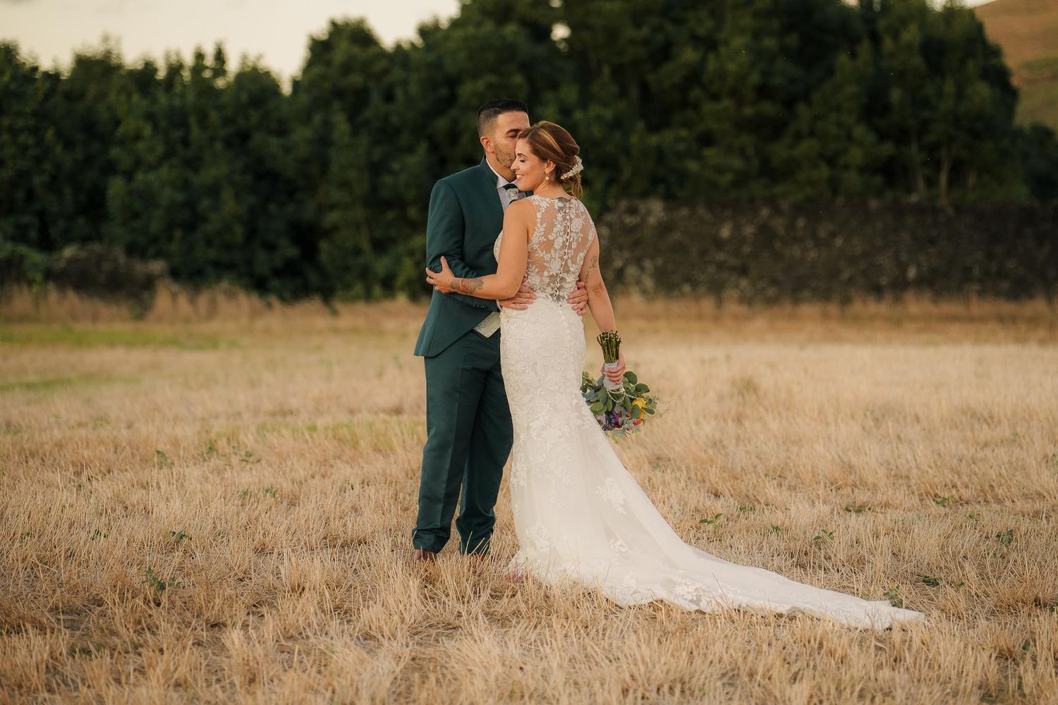 A couple in wedding attire walks through a golden field at sunset holding hands.