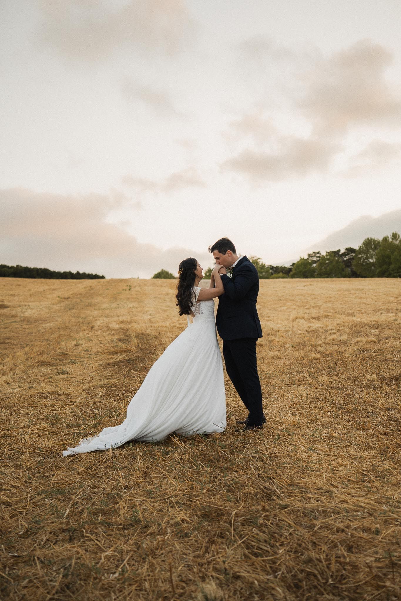 A couple in wedding attire embraces in a golden field at sunset with trees in the background.
