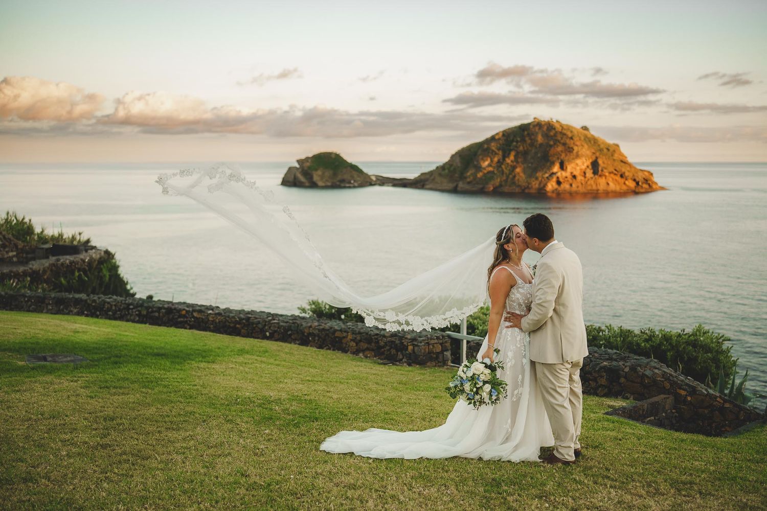 A couple in wedding attire stands on a hilltop overlooking a scenic rocky island during golden hour.