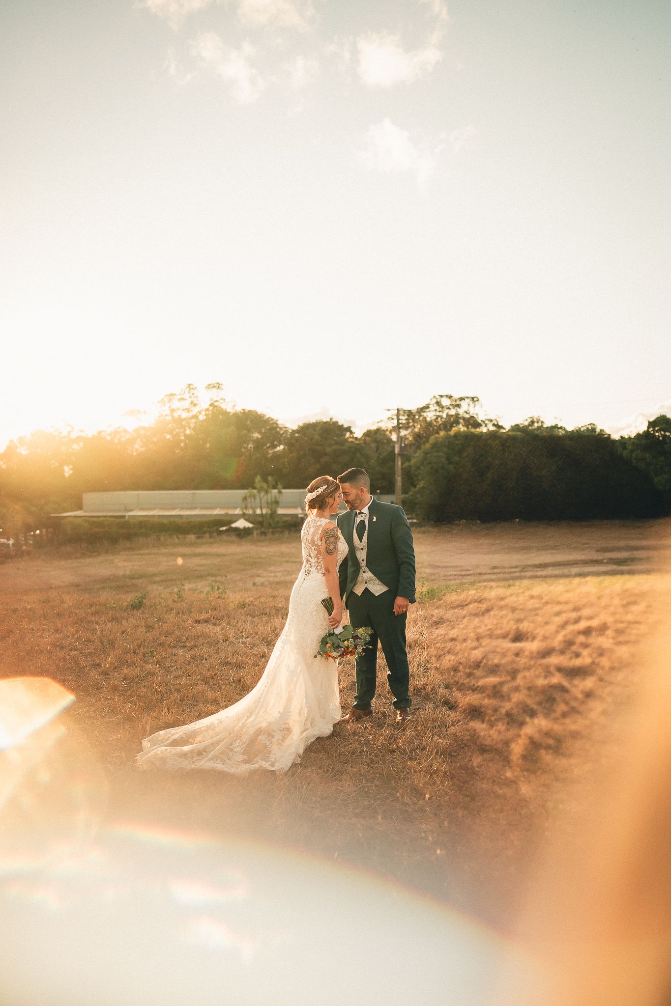 A couple shares a romantic moment in a golden field at sunset while dressed in wedding attire.