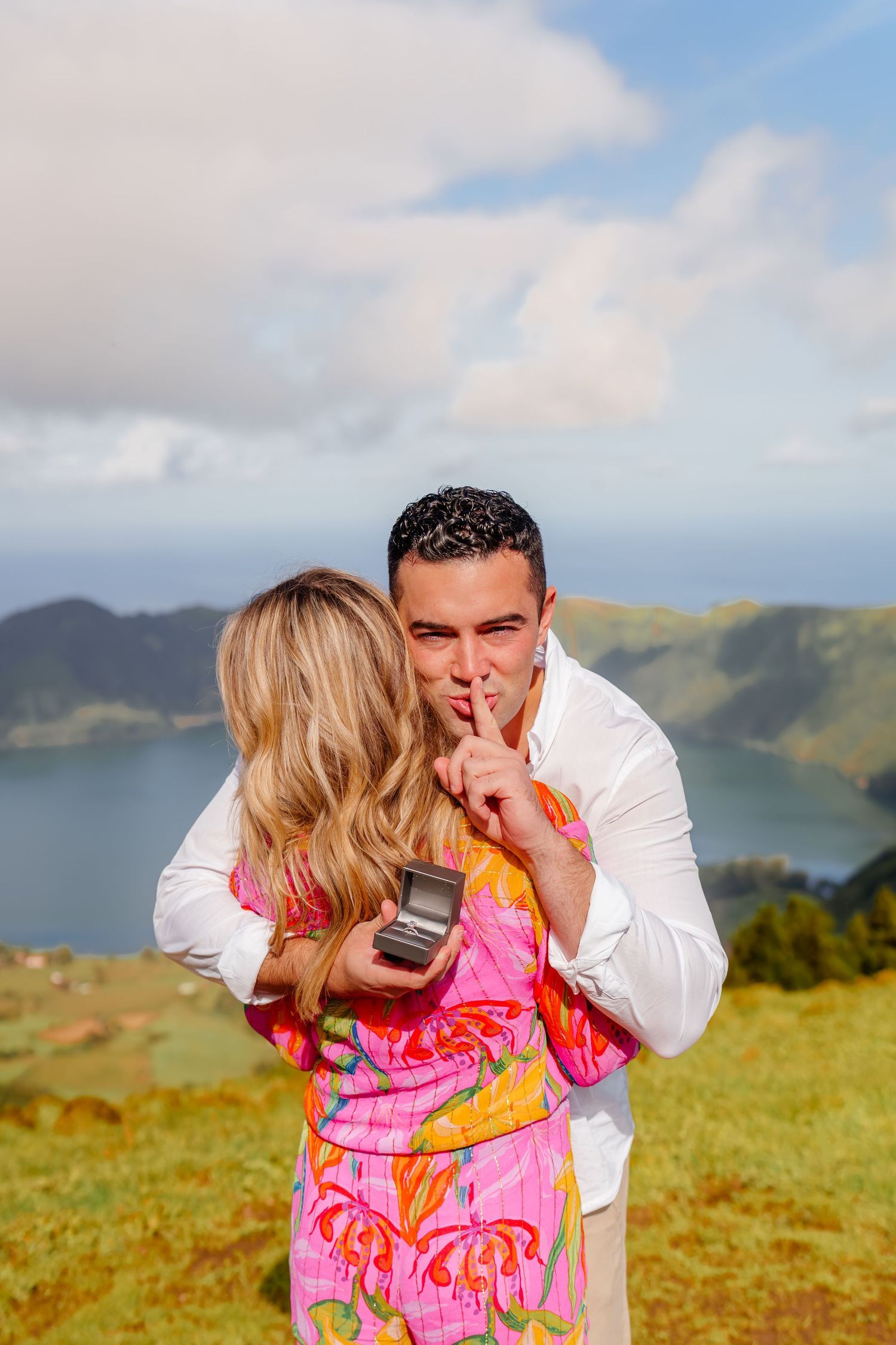 A couple shares a romantic moment on a scenic hillside overlooking mountains and water in bright sunlight.