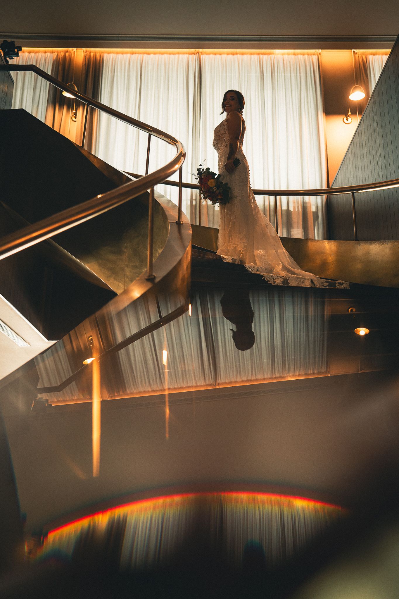 Silhouette of a bride in a flowing white dress stands on a modern staircase with dramatic lighting and reflective surfaces.