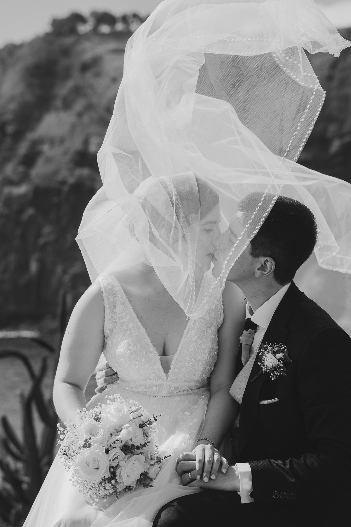Romantic black and white wedding photograph with bride's veil flowing in the wind as couple shares an intimate moment.
