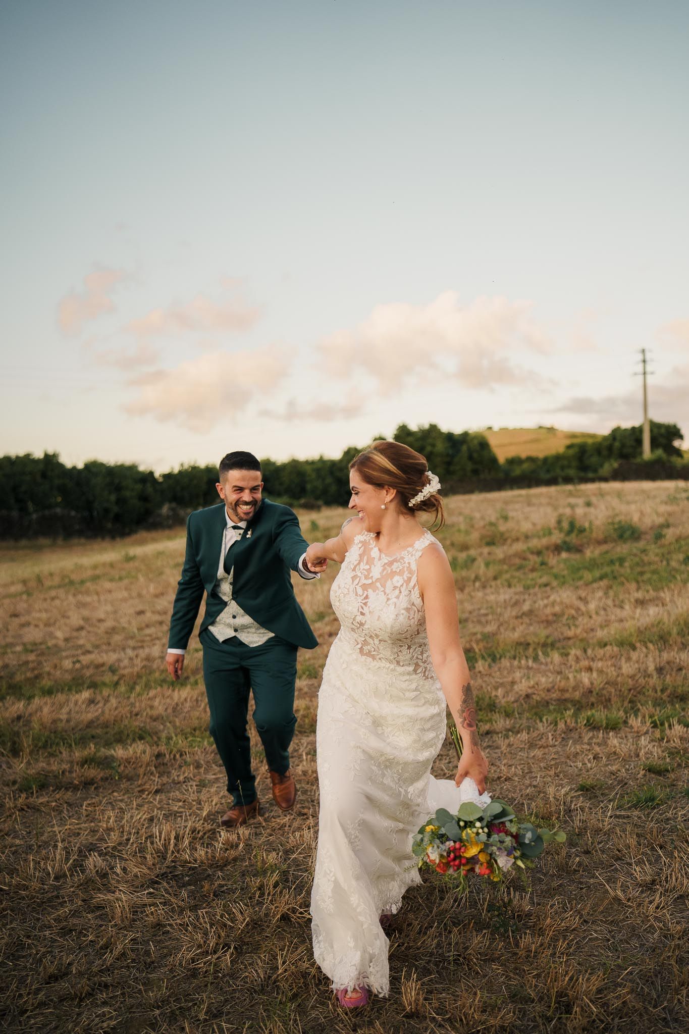 A couple walking through a scenic grassy hillside at sunset in wedding attire holding a colorful bouquet.