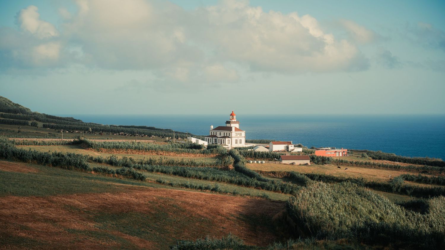 A white lighthouse perches on a coastal cliff overlooking fields and the blue ocean in the Azores.