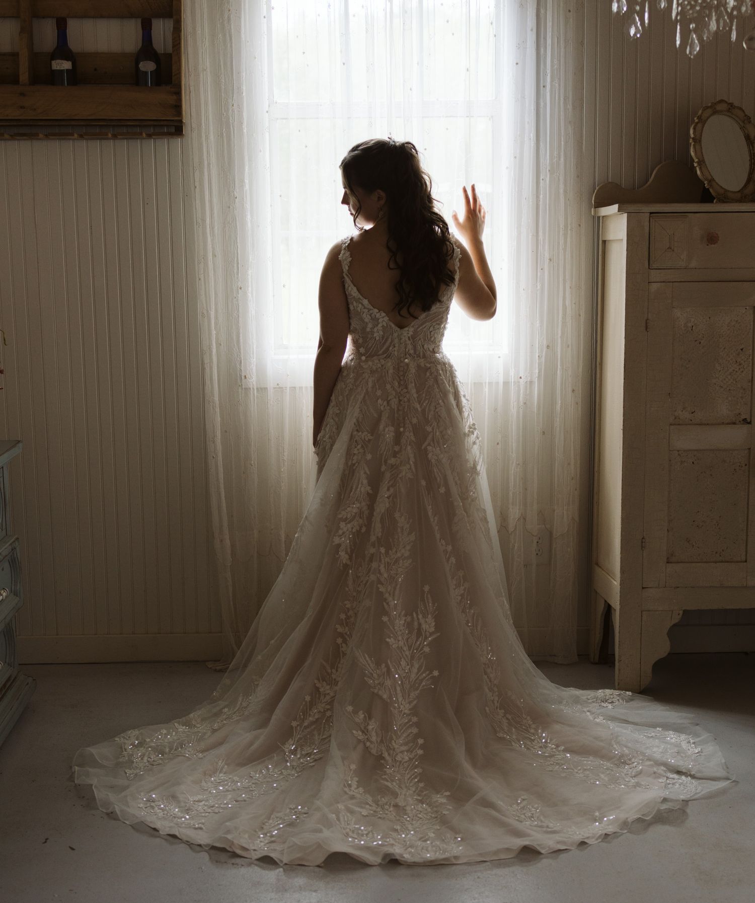 Elegant lace wedding dress with long train photographed in silhouette against bright window.