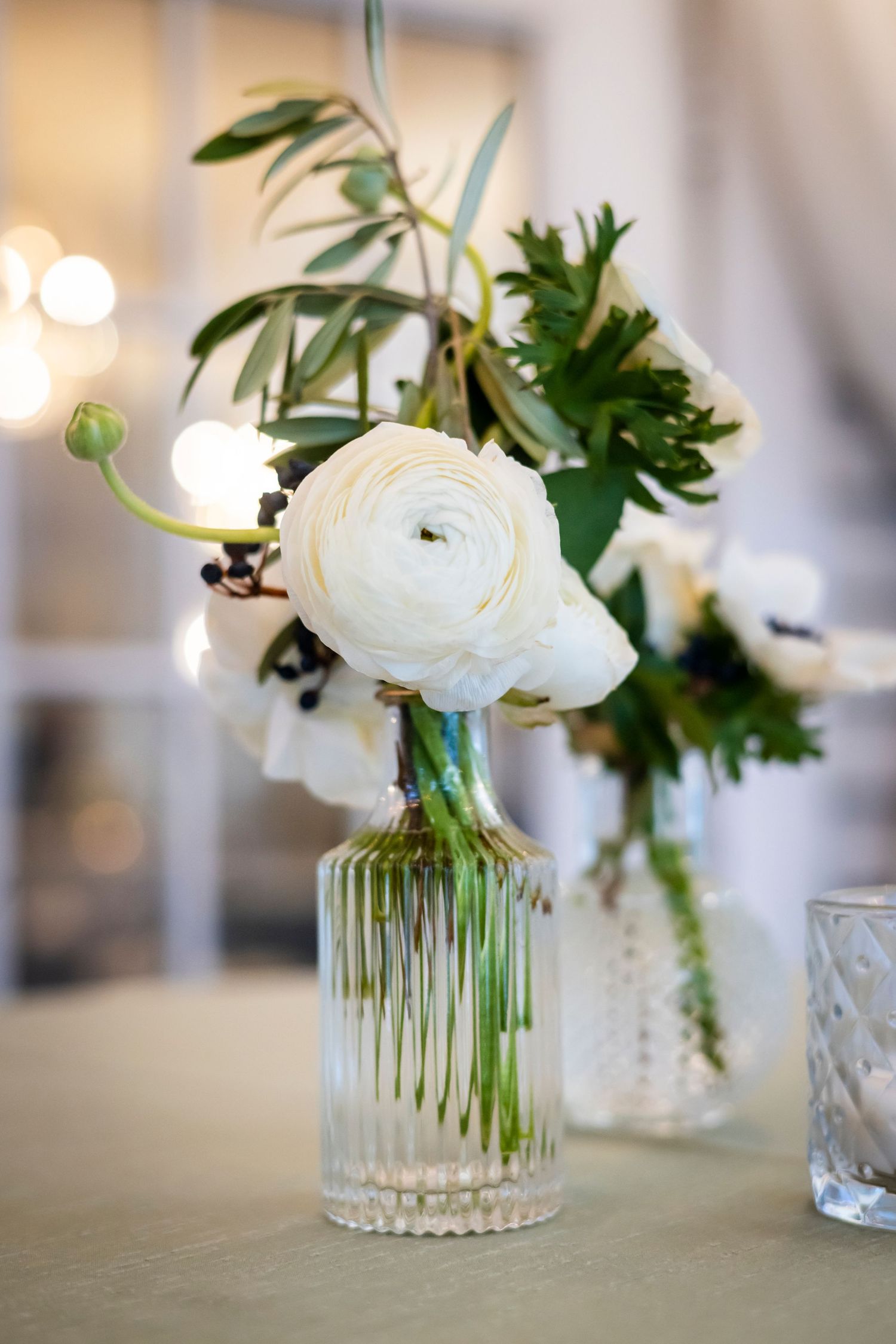 White ranunculus flowers in crystal vases with green foliage as wedding table centerpieces.