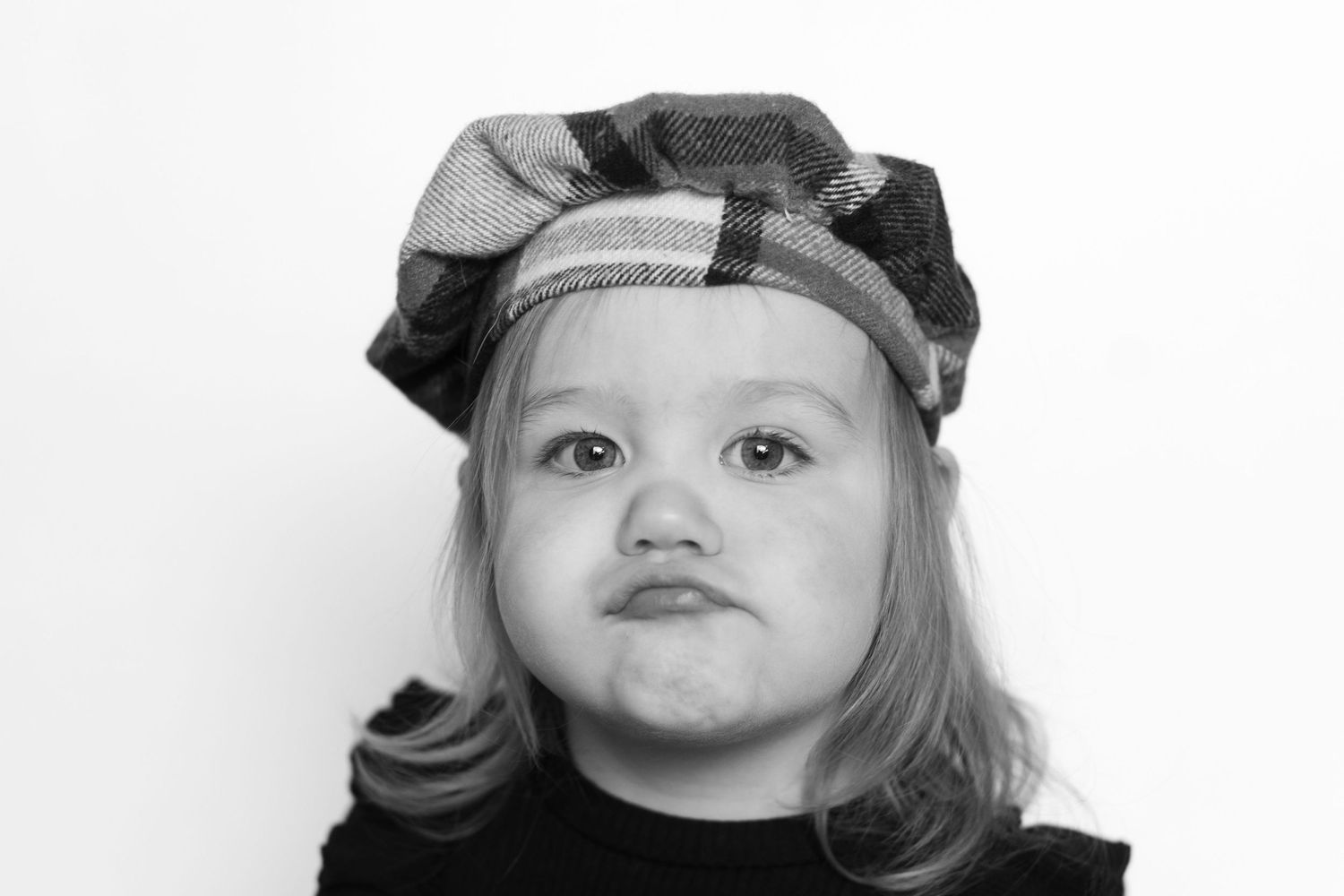 Black and white portrait of a toddler girl wearing a plaid beret, making a pouty face.