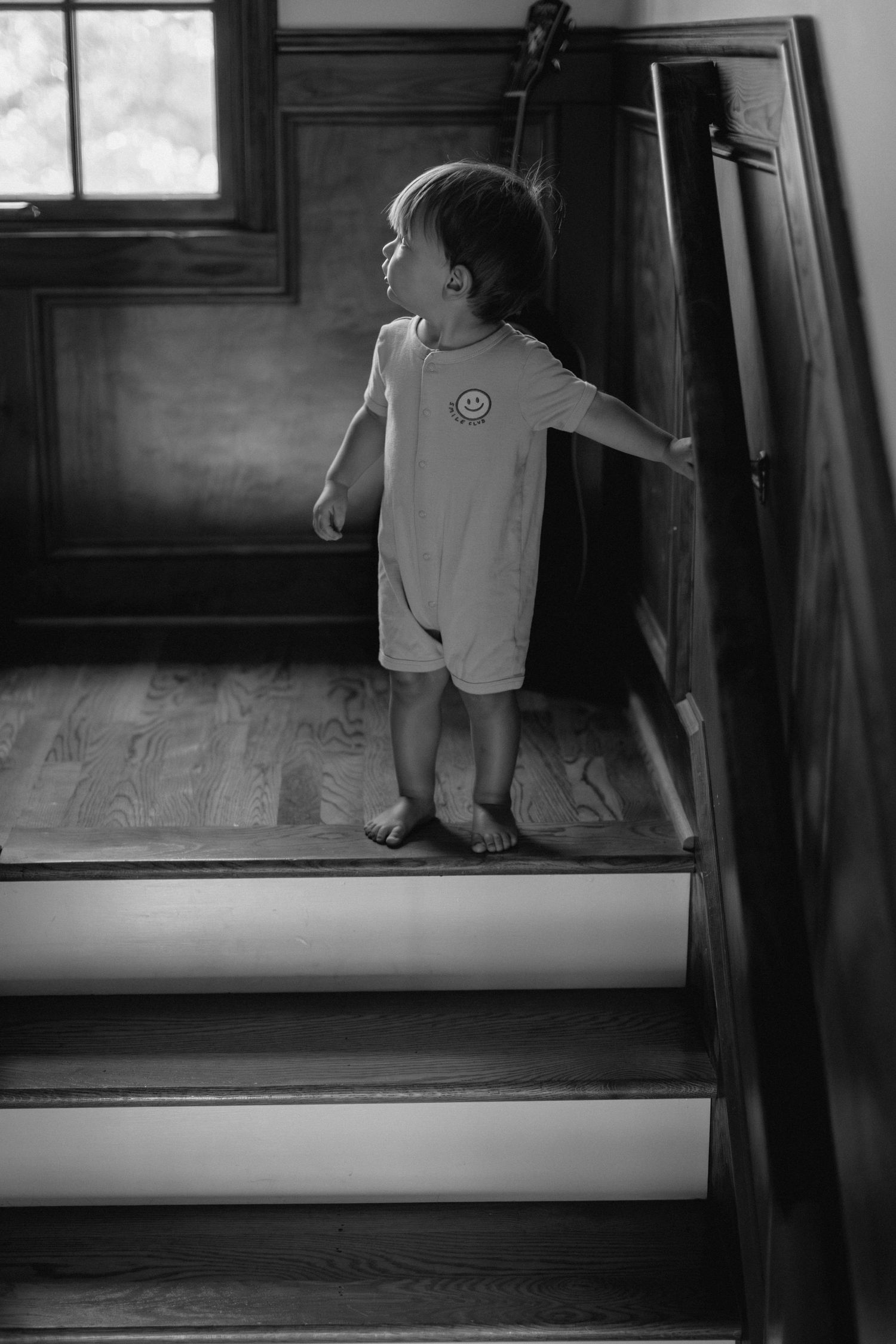 Black and white photo of a toddler in a romper standing on wooden stairs holding the railing.