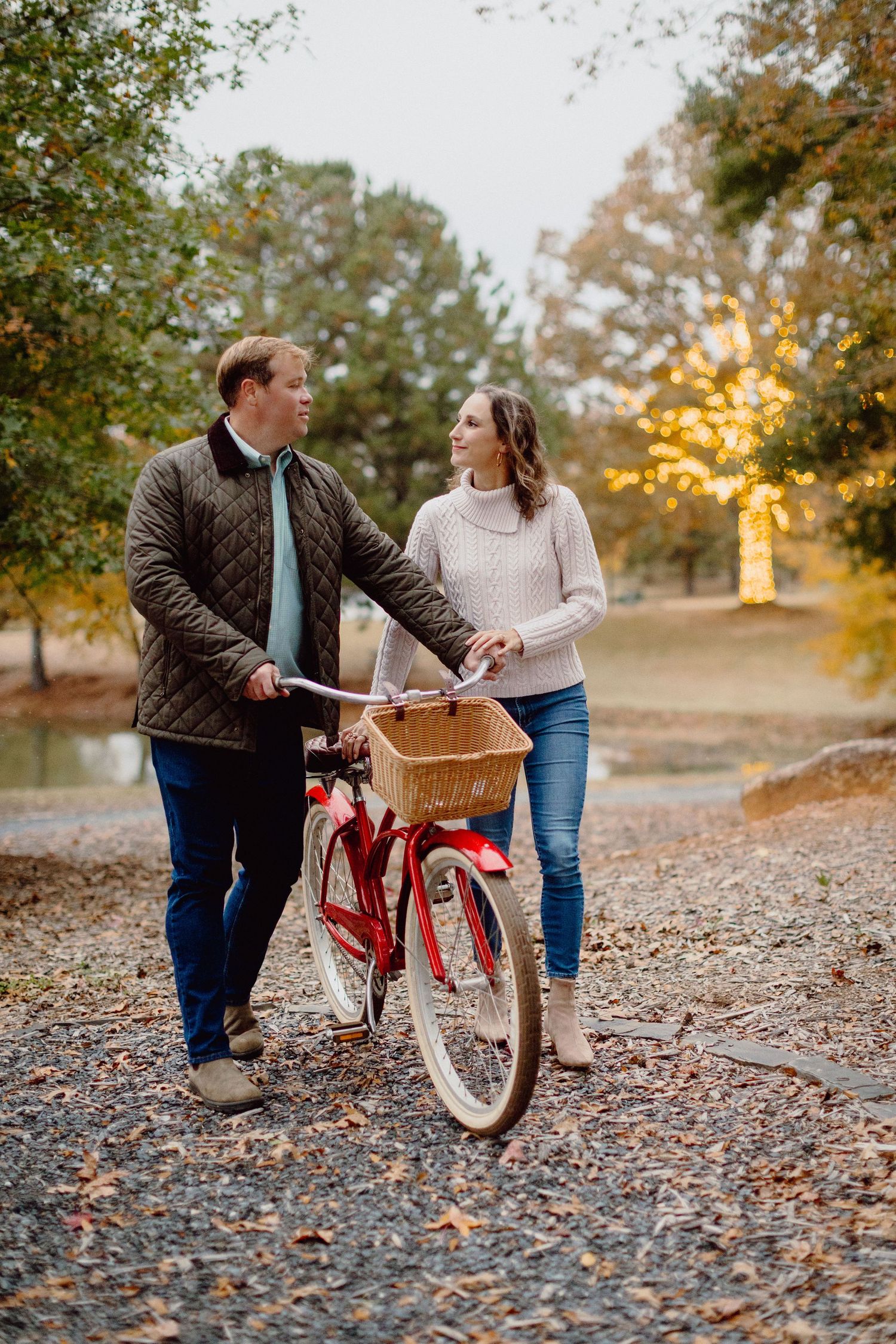 A couple walks together with a vintage red bicycle with basket along a leaf-covered path during autumn sunset.