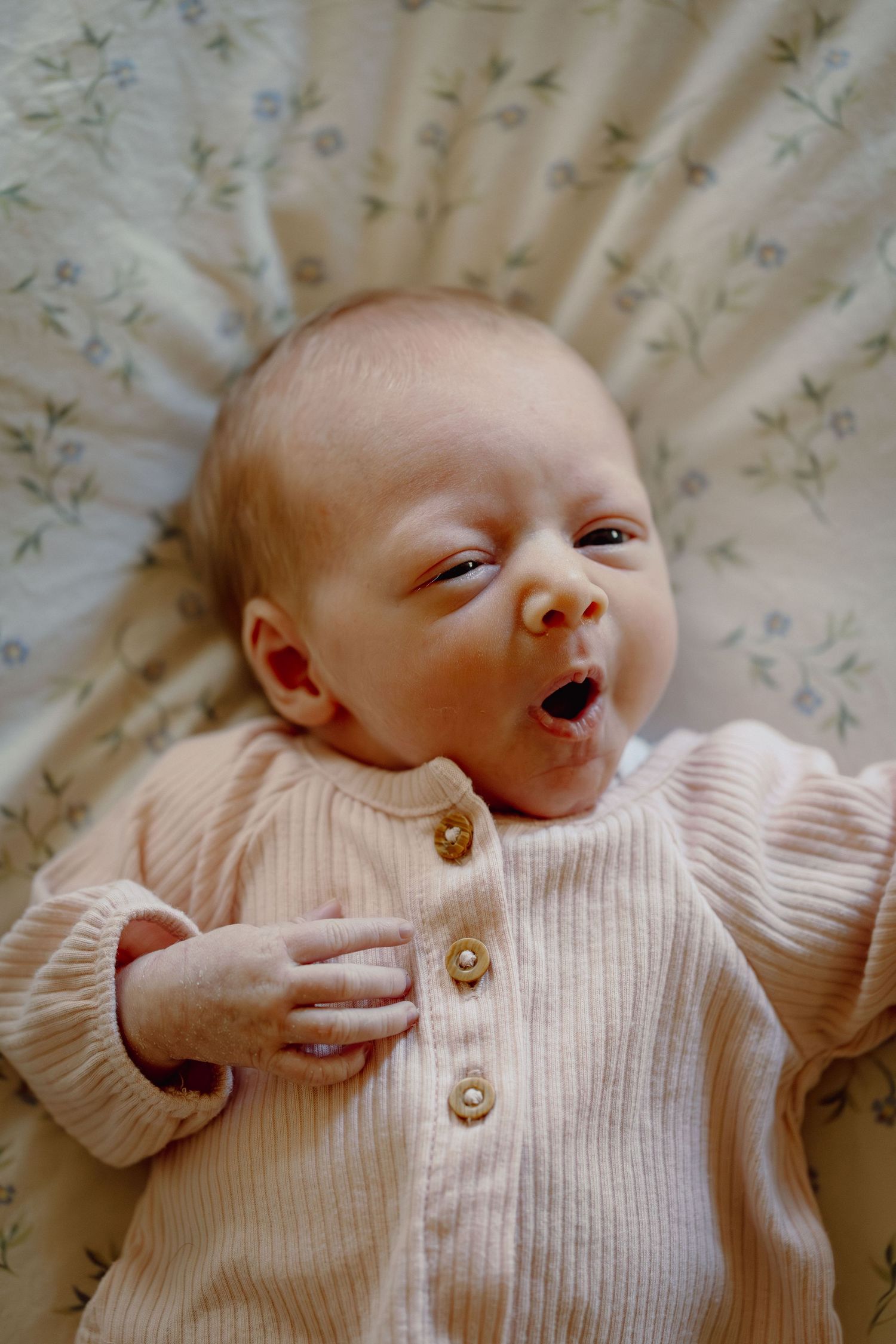 Newborn baby yawning while lying on floral bedding, wearing a ribbed pink button-up onesie.