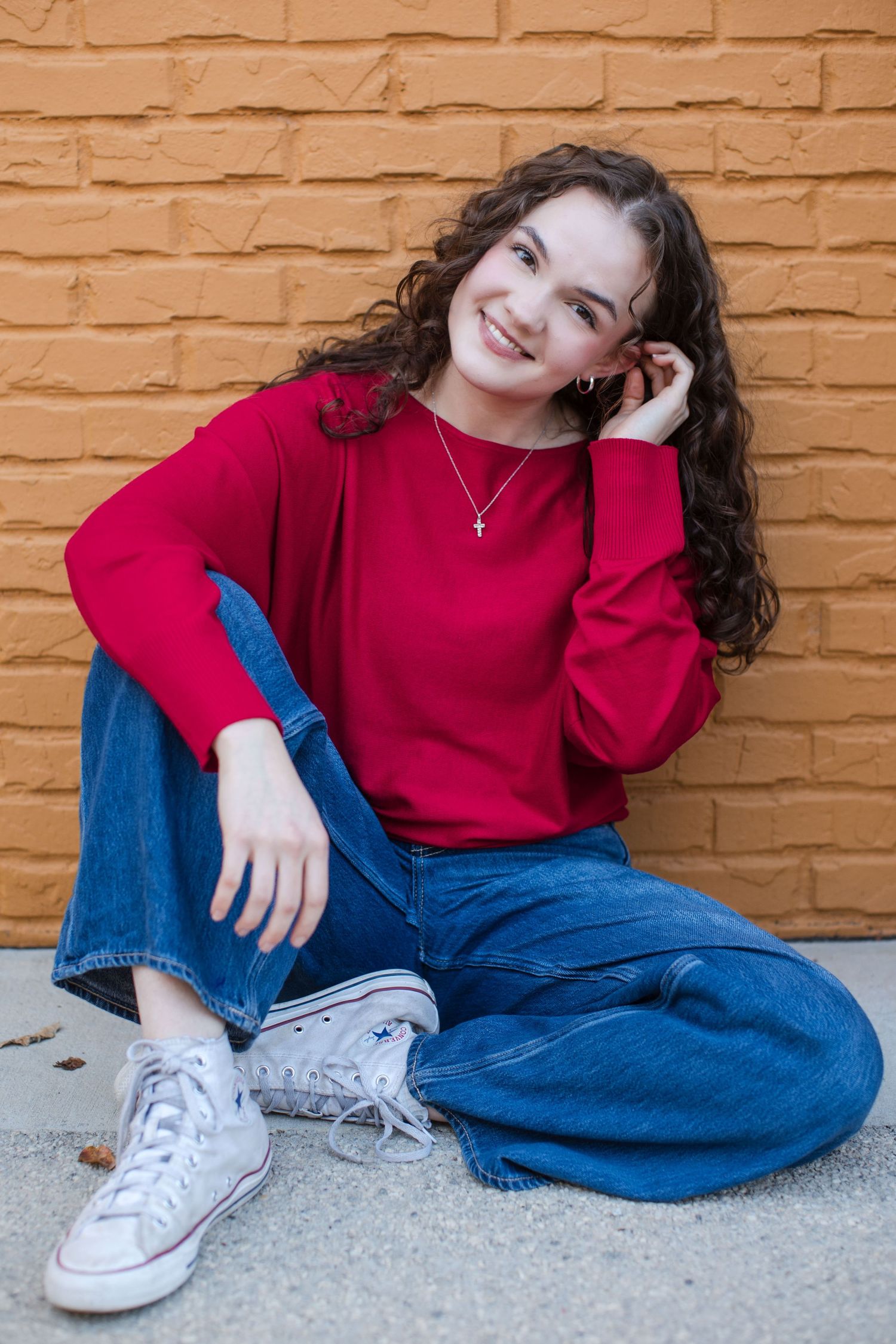 Smiling young woman in red sweater and jeans sitting against orange brick wall wearing white Converse sneakers.