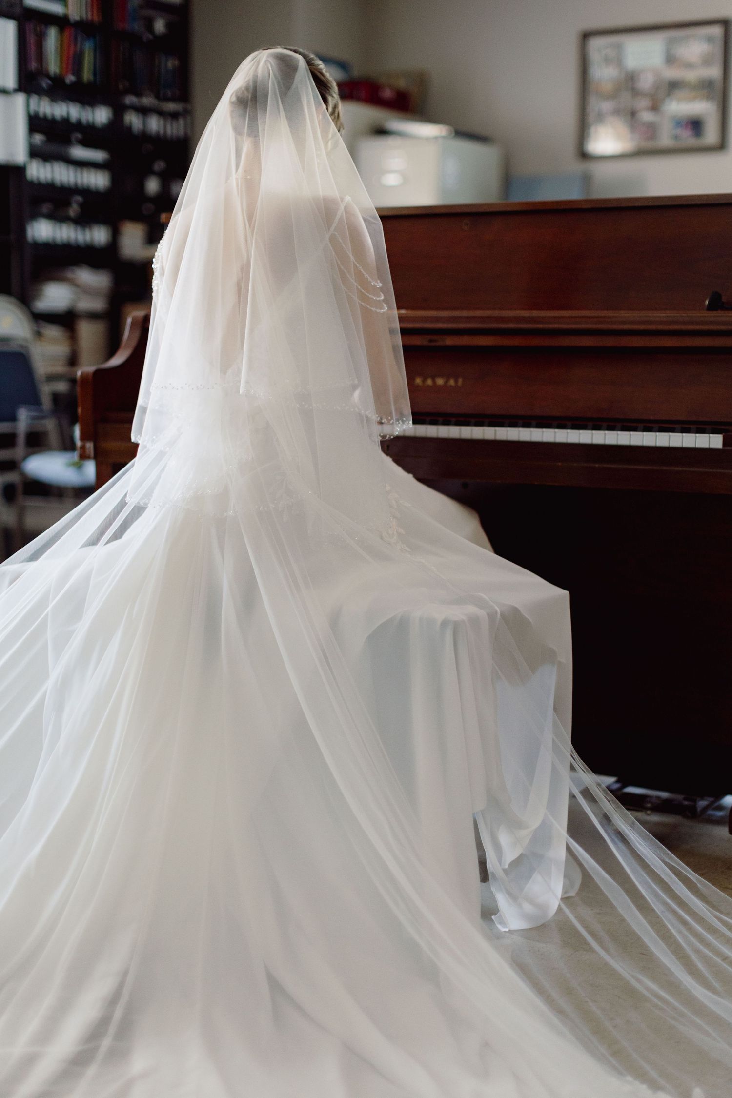 Bride in flowing white wedding gown and veil seated at a Kawai upright piano in an elegant room.