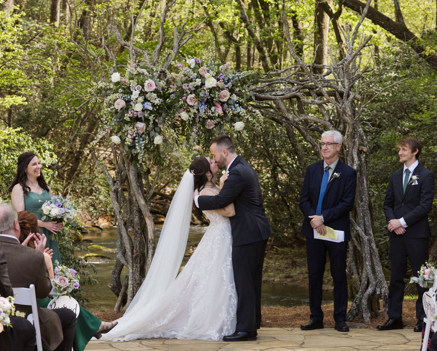 Outdoor wedding ceremony taking place among trees with guests seated on white chairs.