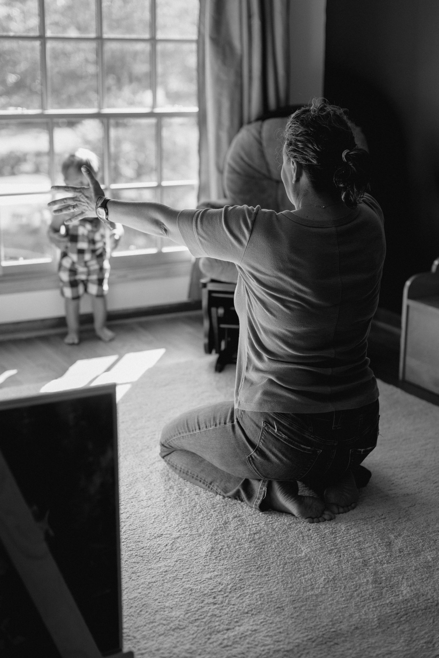 Black and white photo of a mother kneeling on carpet, reaching toward her toddler standing by a sunlit window indoors.