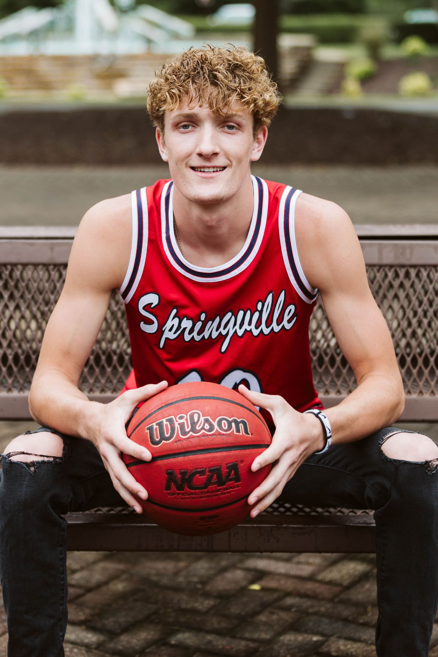 Young man in Springville red basketball jersey holding a Wilson NCAA basketball while sitting on a bench outdoors.