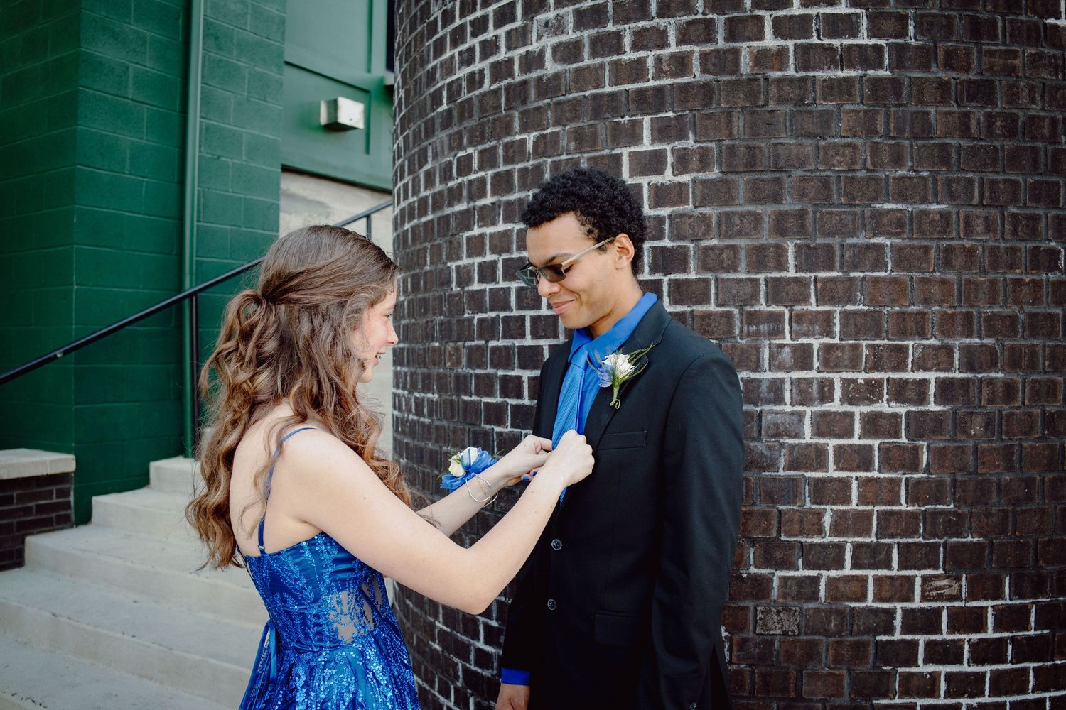 A young woman in a blue sequin dress pins a boutonniere on a young man in a black suit at prom.