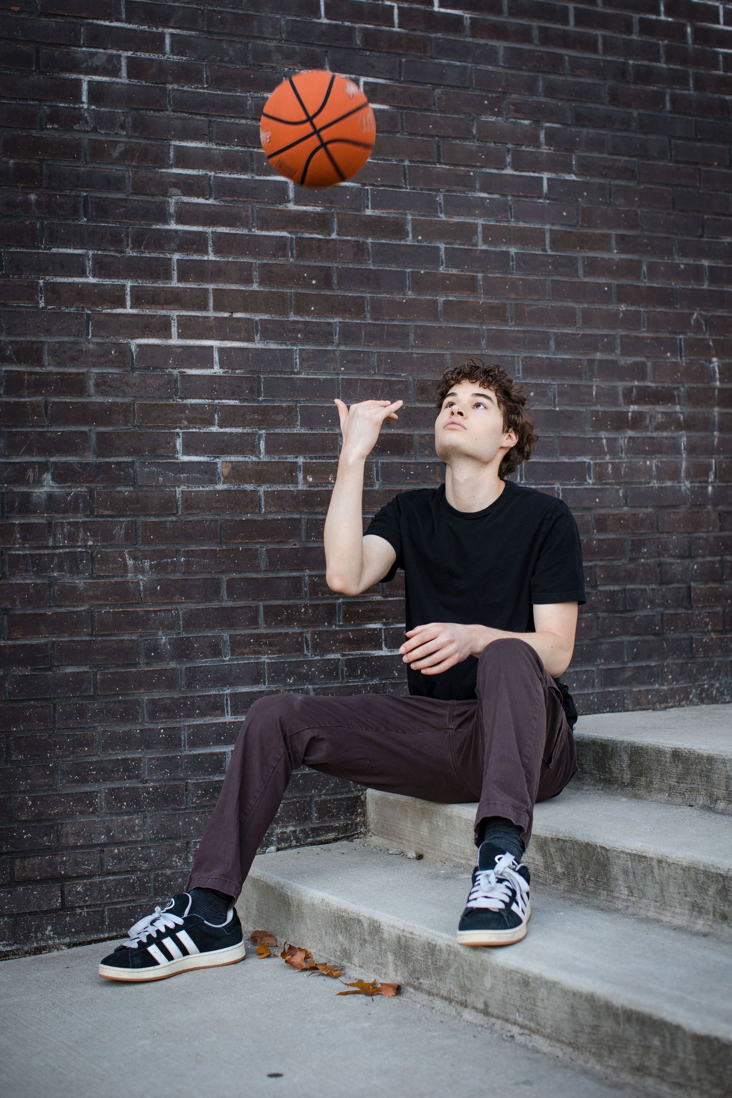 A person in black shirt and brown pants performs a creative basketball trick while sitting on concrete steps.