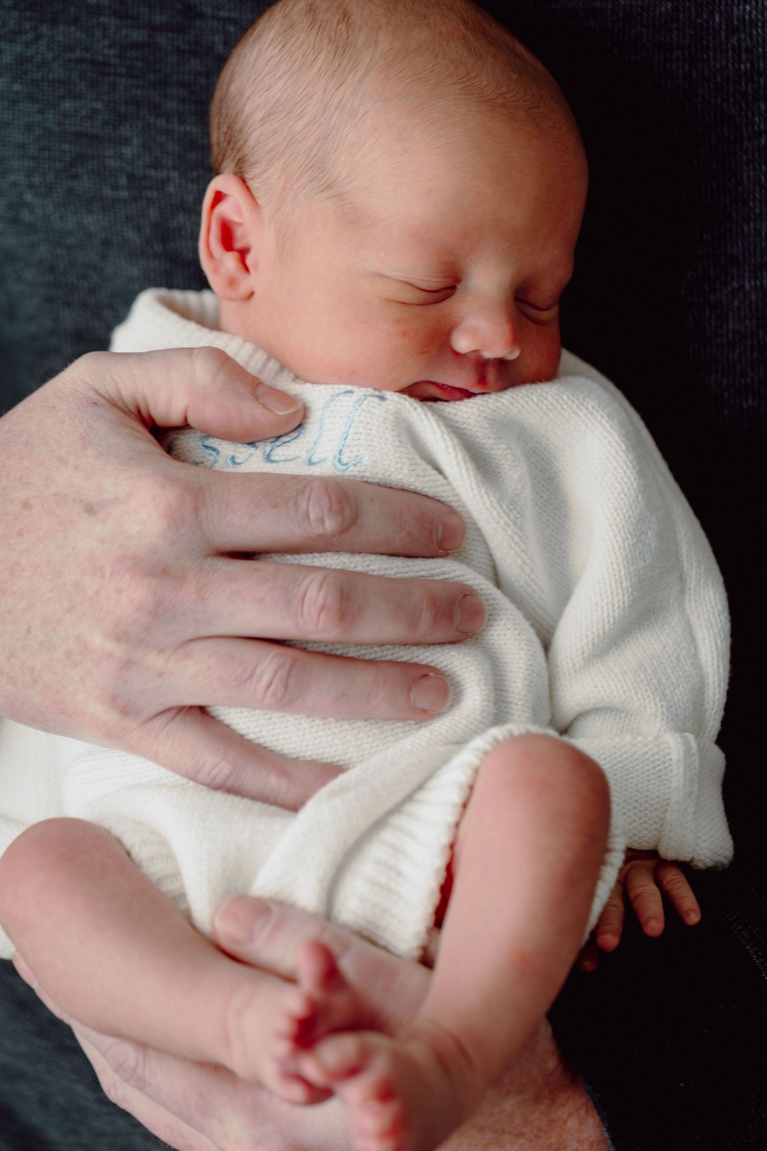 A sleeping newborn baby wearing a white knit outfit is cradled in protective hands against a dark background.