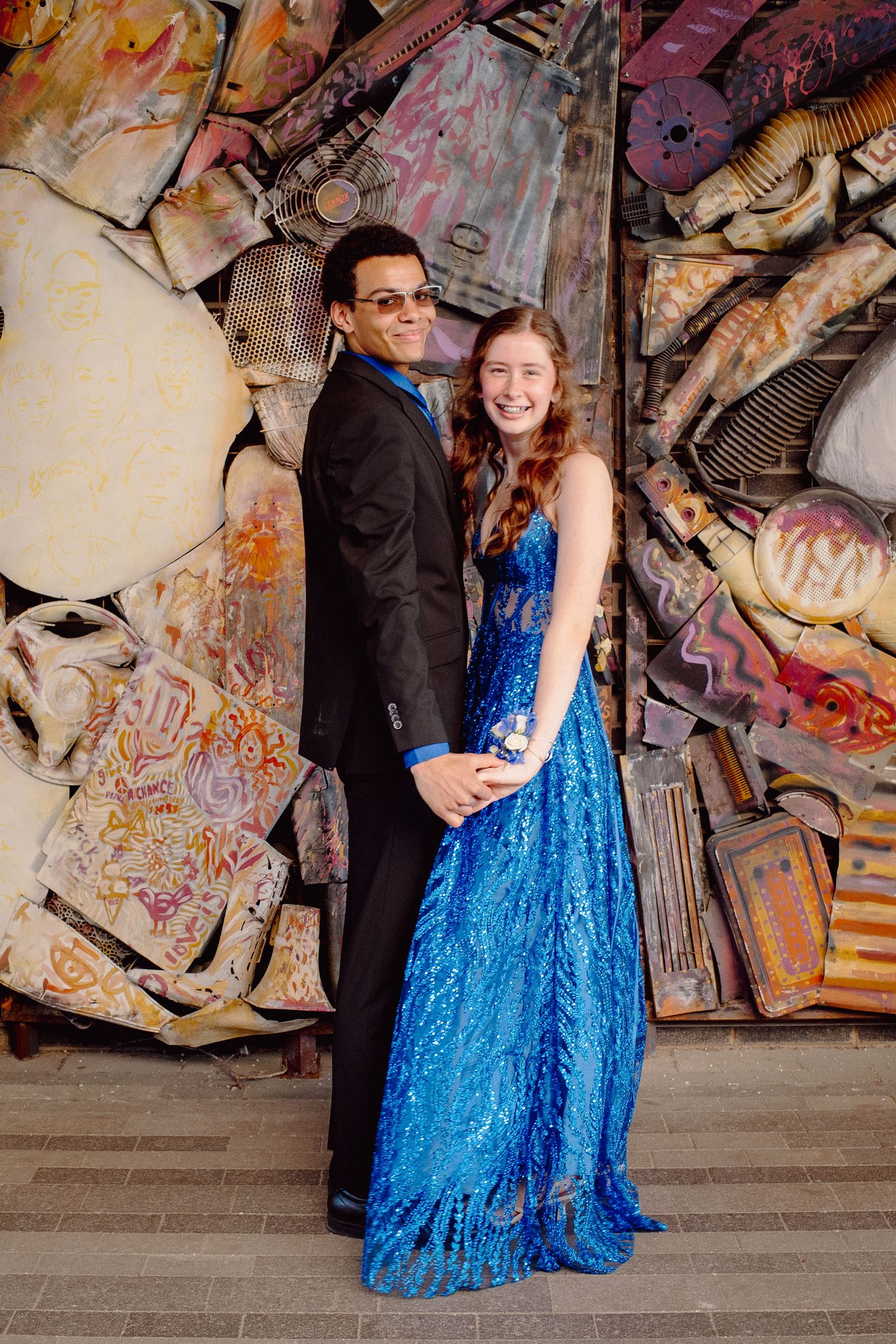 Teen couple in prom attire, girl in blue sequin gown and boy in black suit, posing before colorful mixed-media wall art.